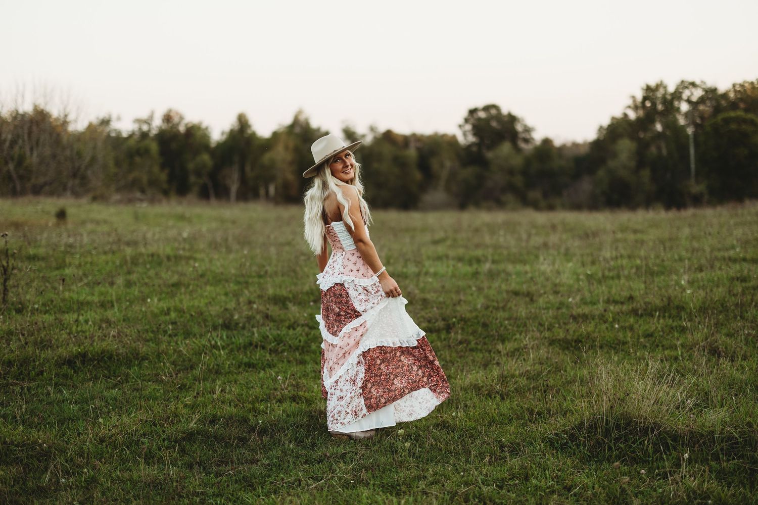 Woman in floral maxi dress and white hat twirling in green field at golden hour.
