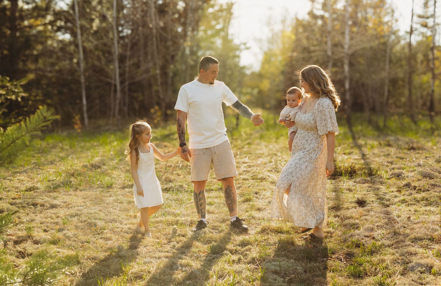 Family of four walking in sunny forest meadow, parents holding hands with young daughters in white dresses.