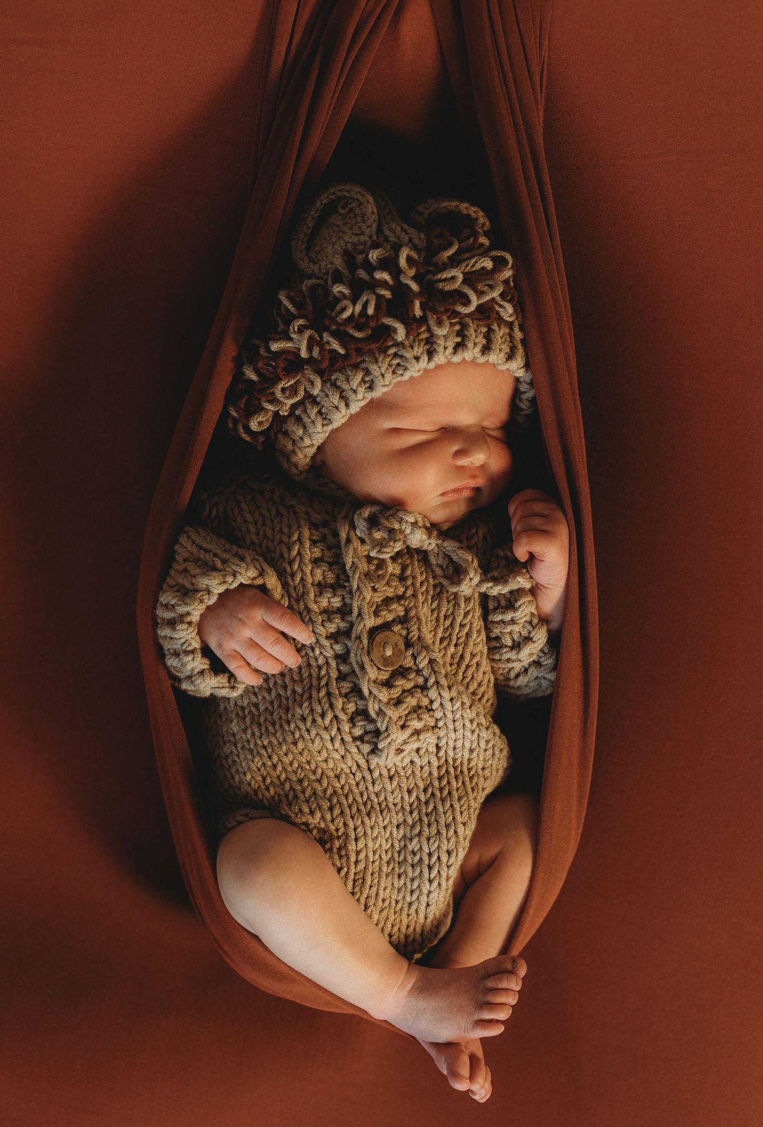 Sleeping newborn baby in knit bear hat and sweater outfit, nestled in a rust-colored fabric hammock.