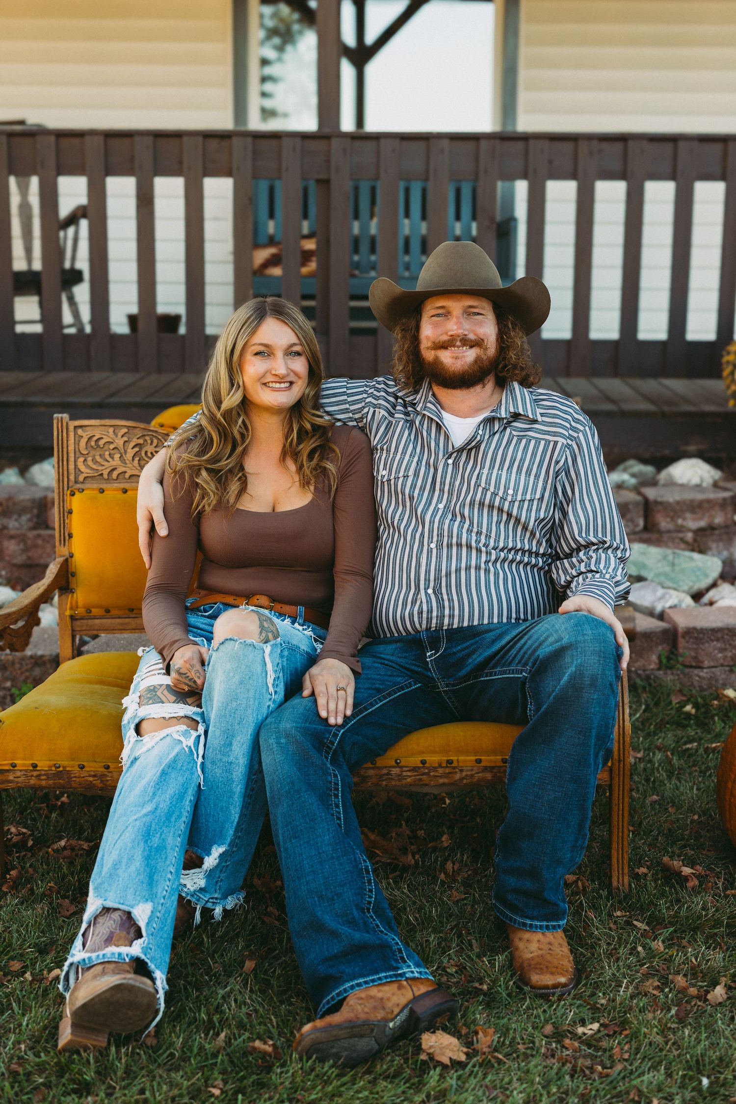 Couple sitting together on rustic wooden bench in autumn yard wearing casual denim and boots.