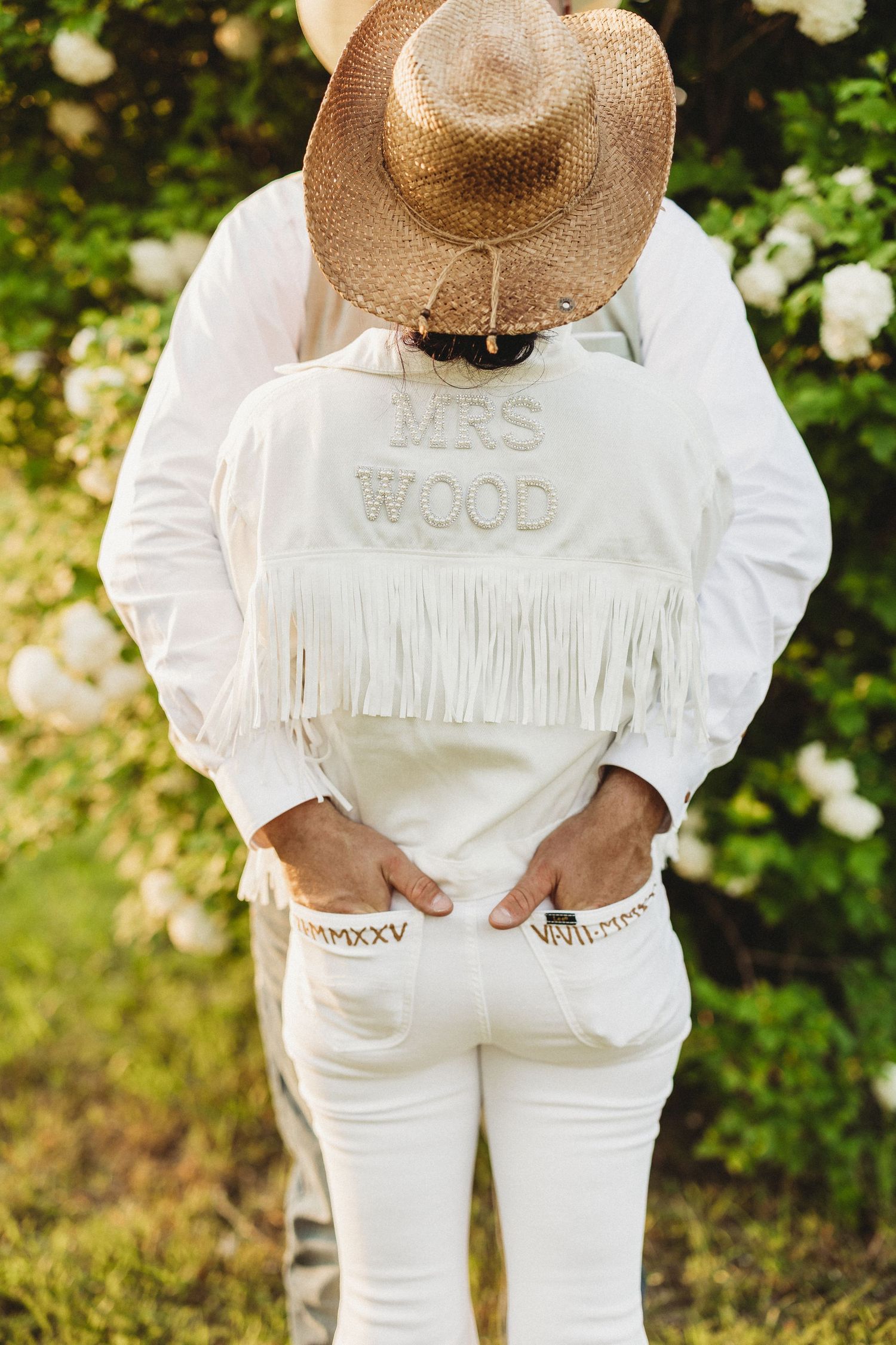 Person wearing all white clothing and a straw hat stands in front of blooming flowers.