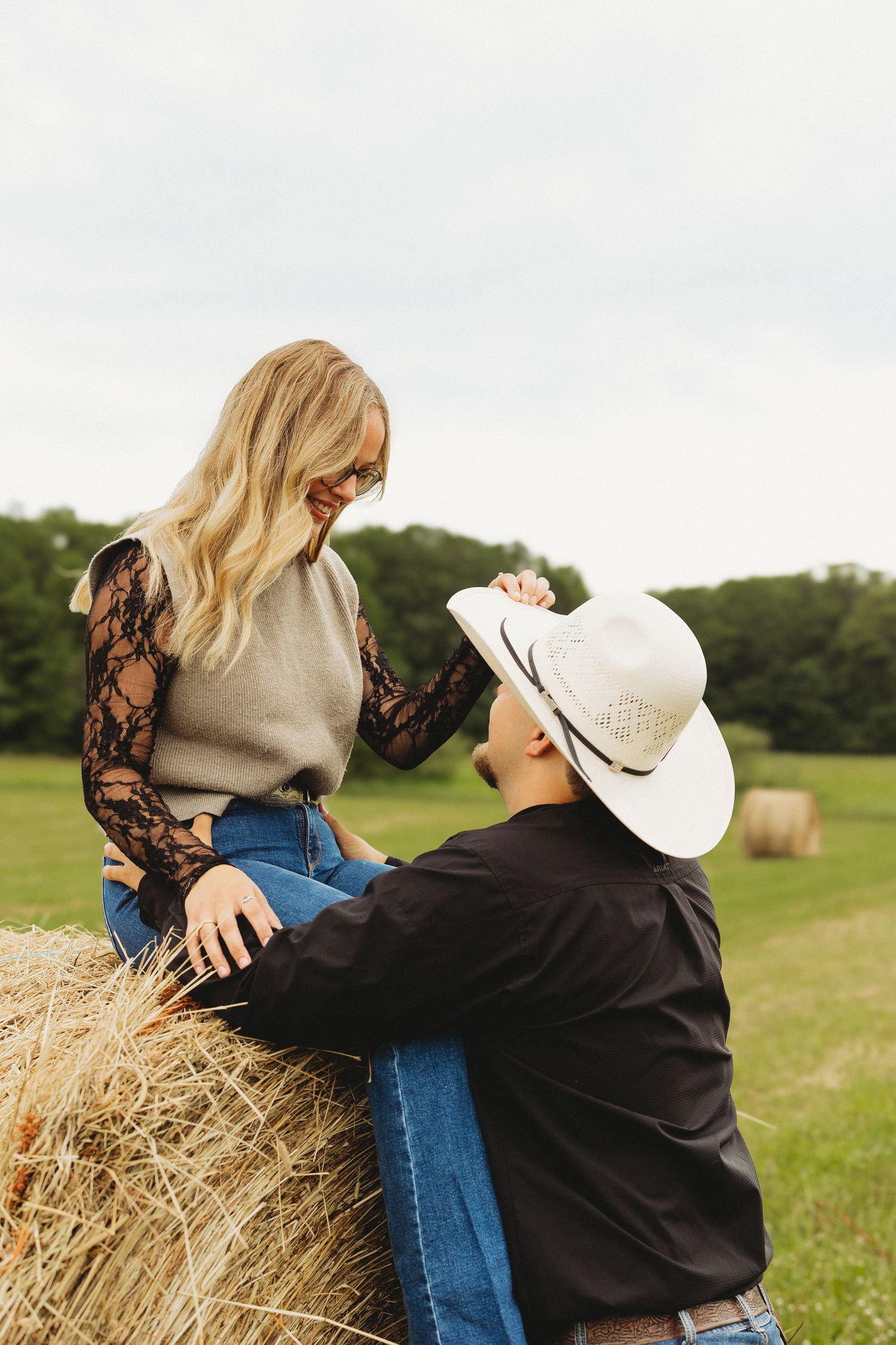 Couple in a field with hay bales, woman sitting on bale in lace top, man in cowboy hat reaching up to her.