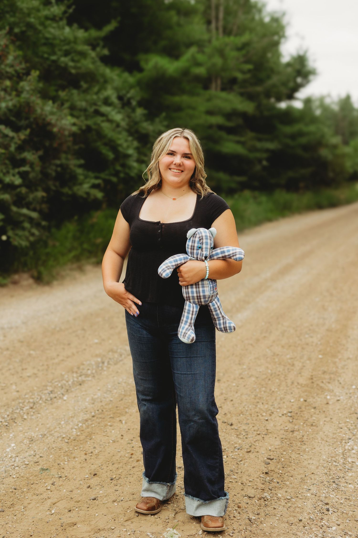 Smiling blonde woman in black top and jeans holding plaid teddy bear on dirt road.