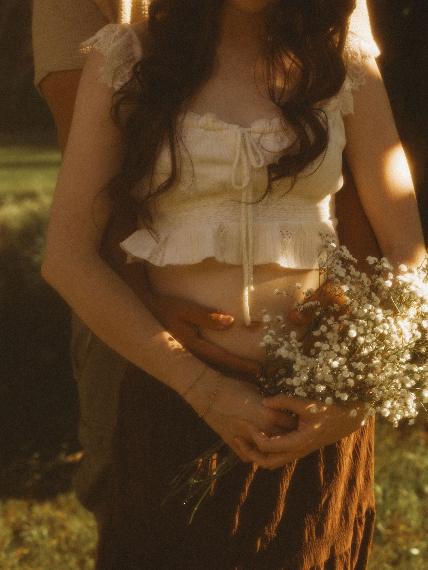 Young woman in white ruffled crop top holding baby's breath wildflowers in golden sunlight outdoors.