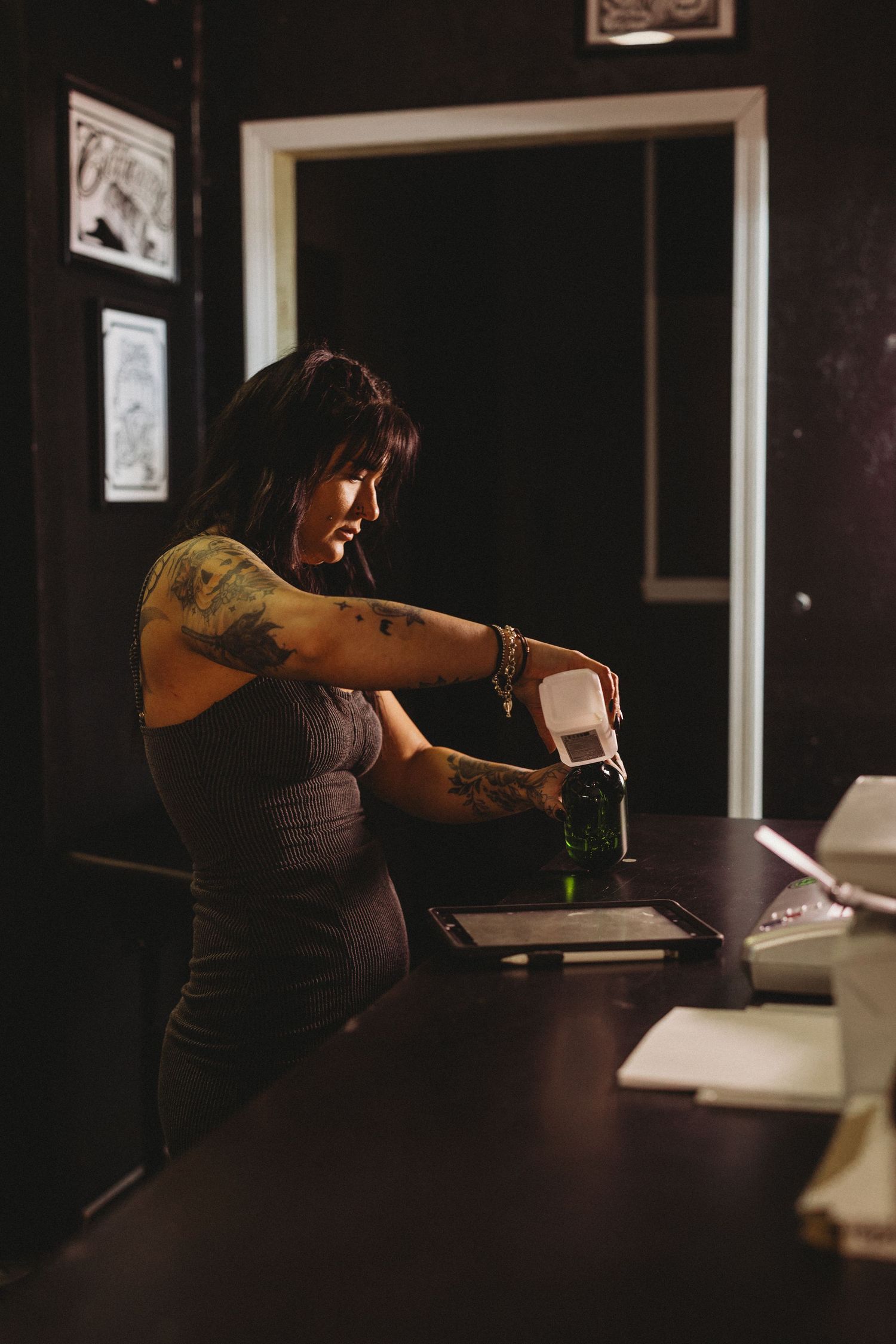 Tattooed woman working at a dark tattoo studio desk, pouring liquid into a container with artwork on walls.