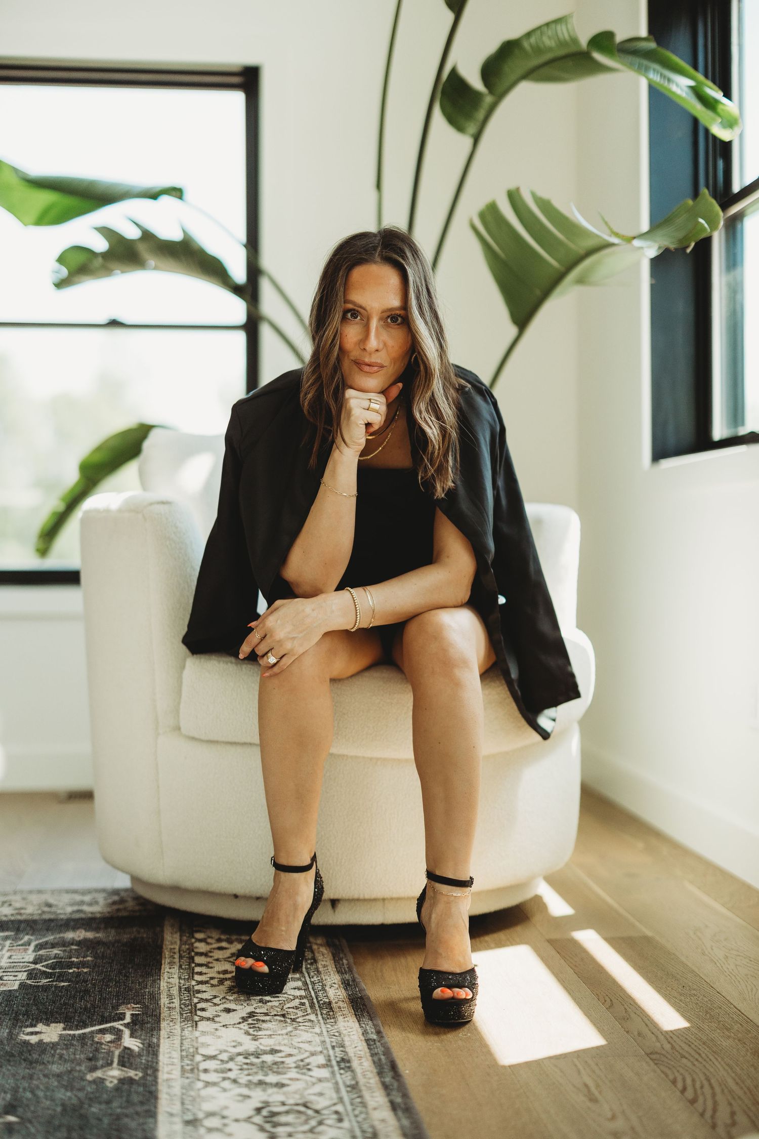 Woman in black blazer and heels sitting in white armchair with tropical plant backdrop in modern interior.
