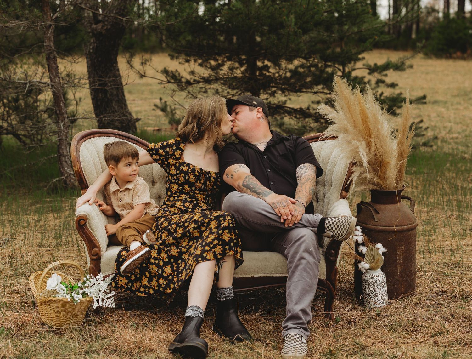A couple kisses on a vintage sofa outdoors while their young son sits beside them in a rustic field setting.