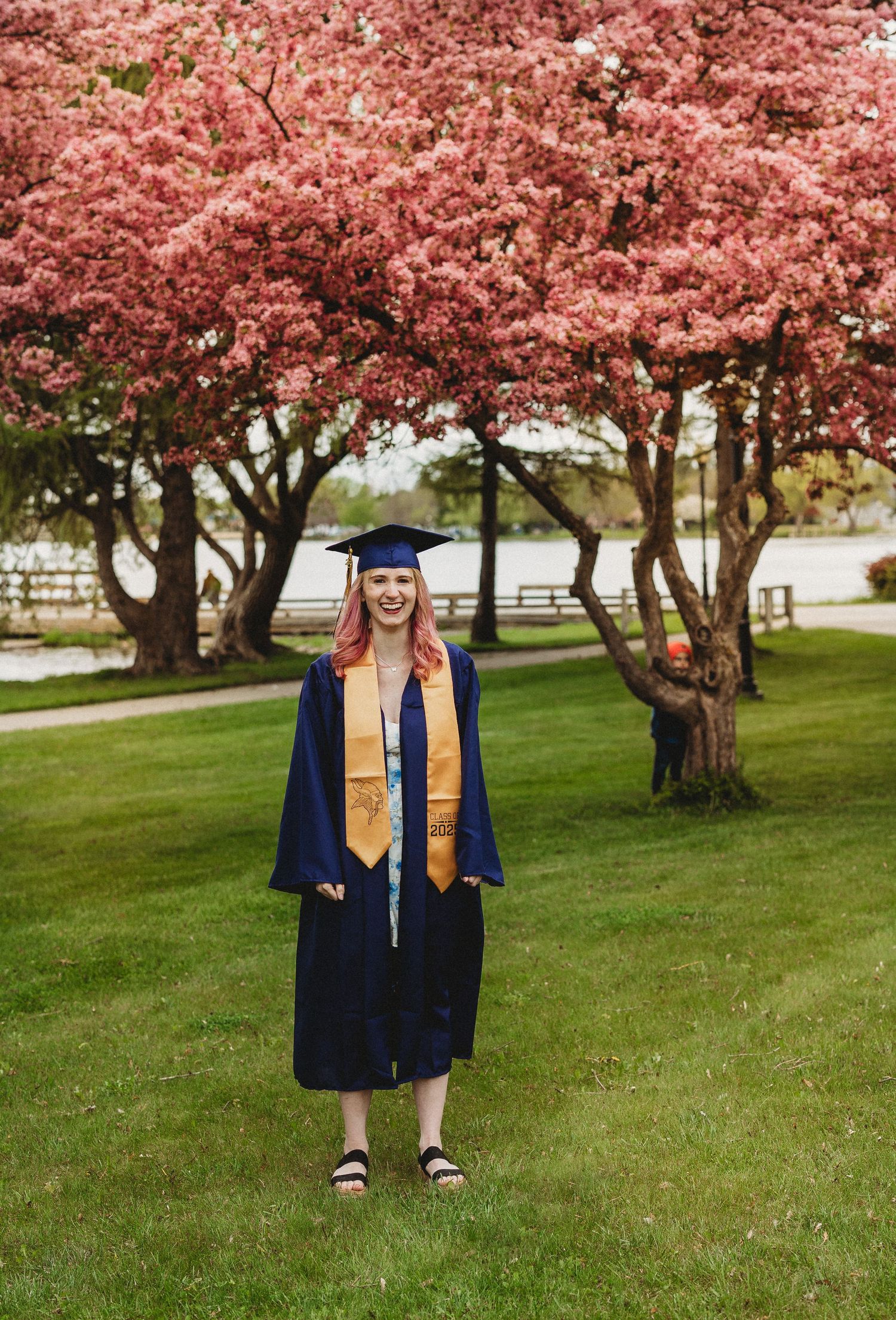 Graduate in traditional blue cap and gown stands beneath pink flowering cherry trees.