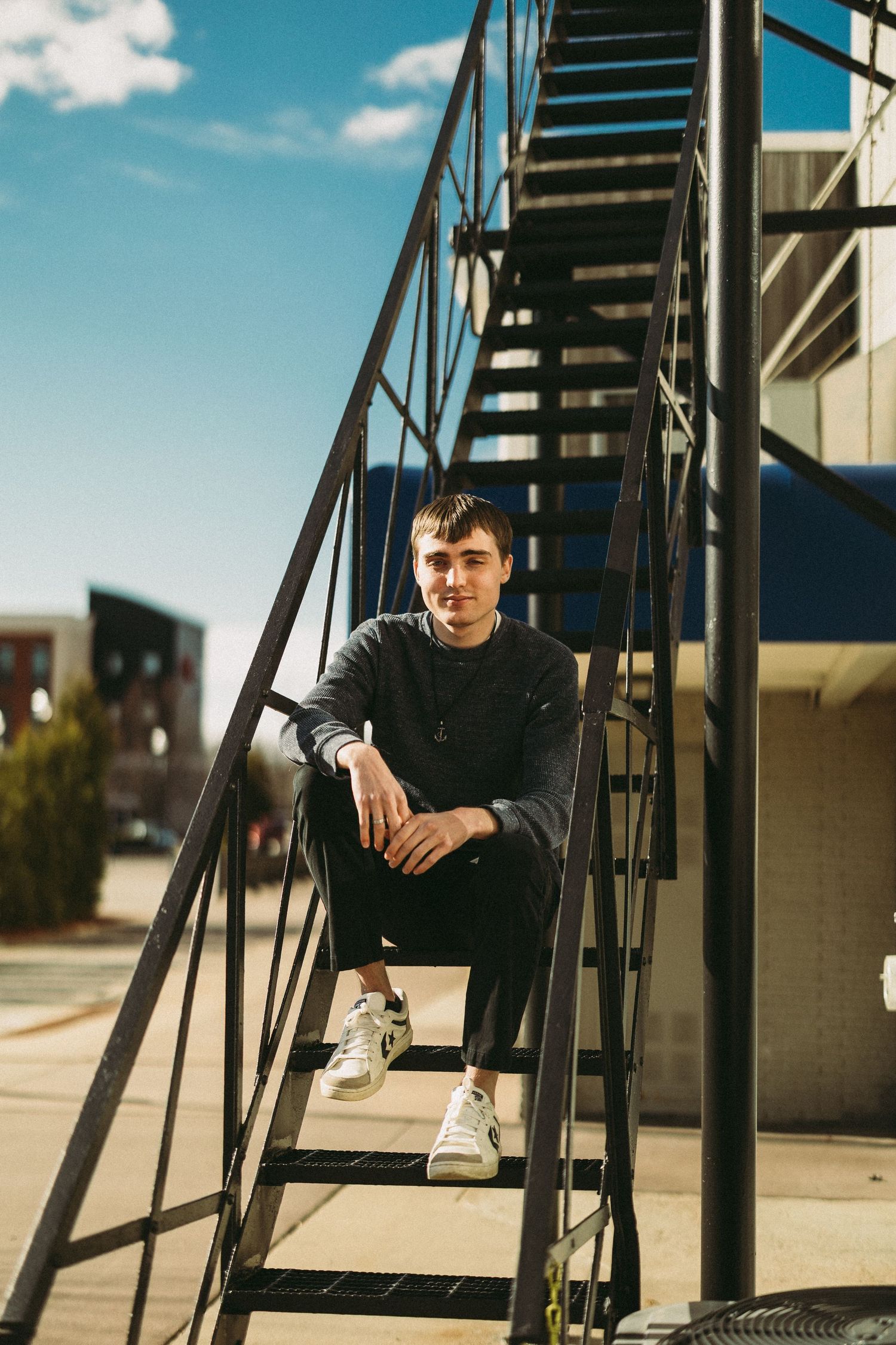 Person in dark clothing sits casually on outdoor metal staircase against blue sky.