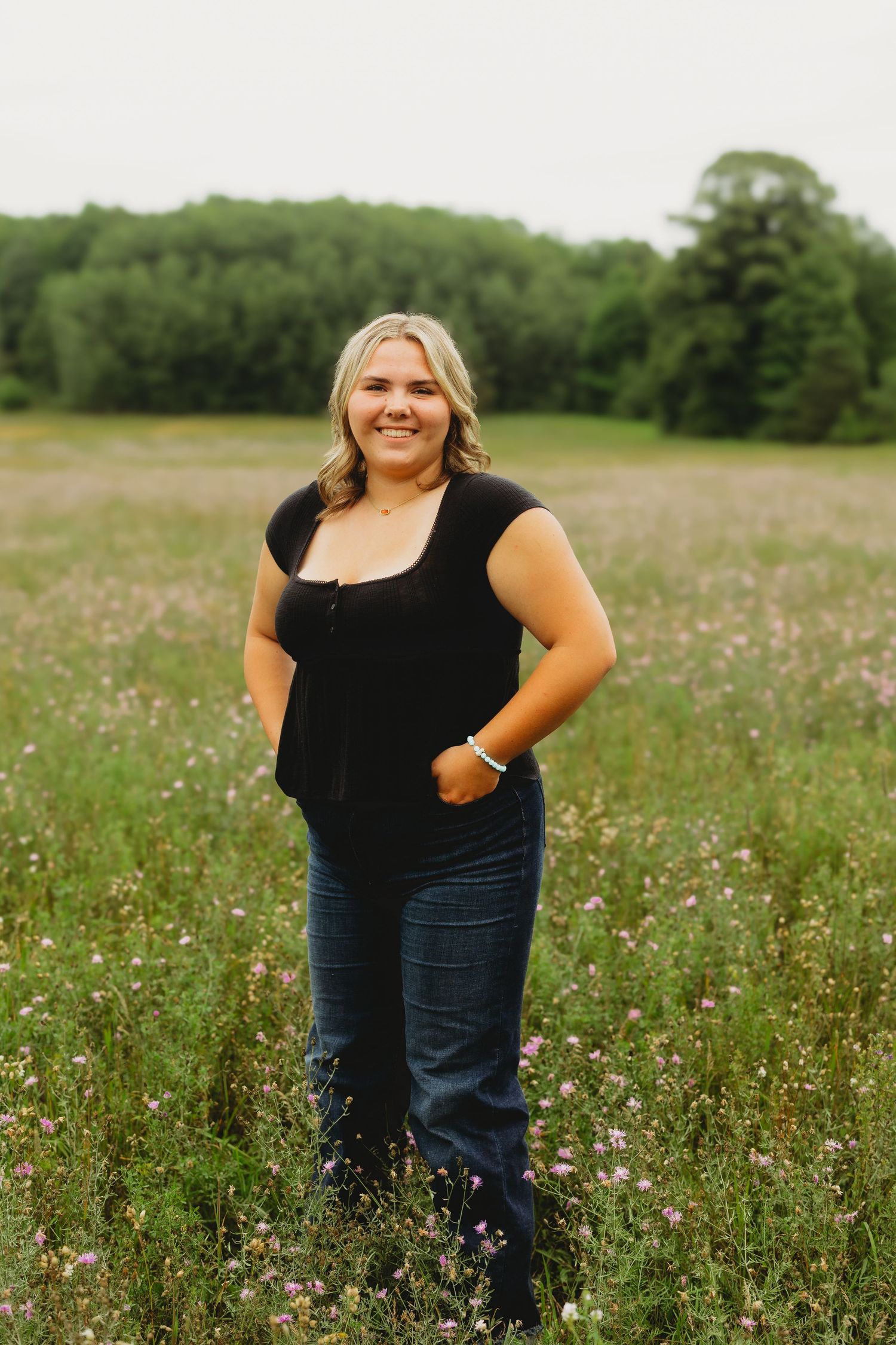 Young blonde woman smiling in a wildflower meadow, wearing a black top and jeans, with green trees in the background.