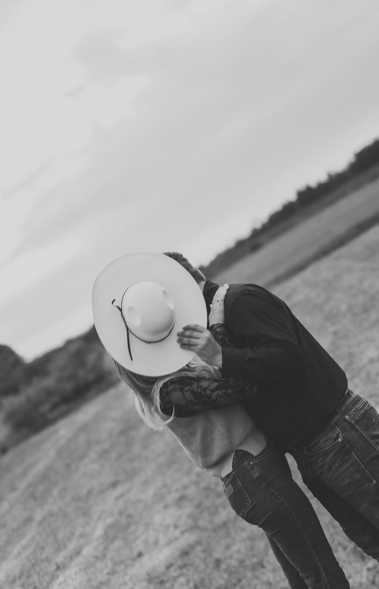 Black and white photo of a couple kissing outdoors, faces hidden by a wide-brim cowboy hat, in an open field.