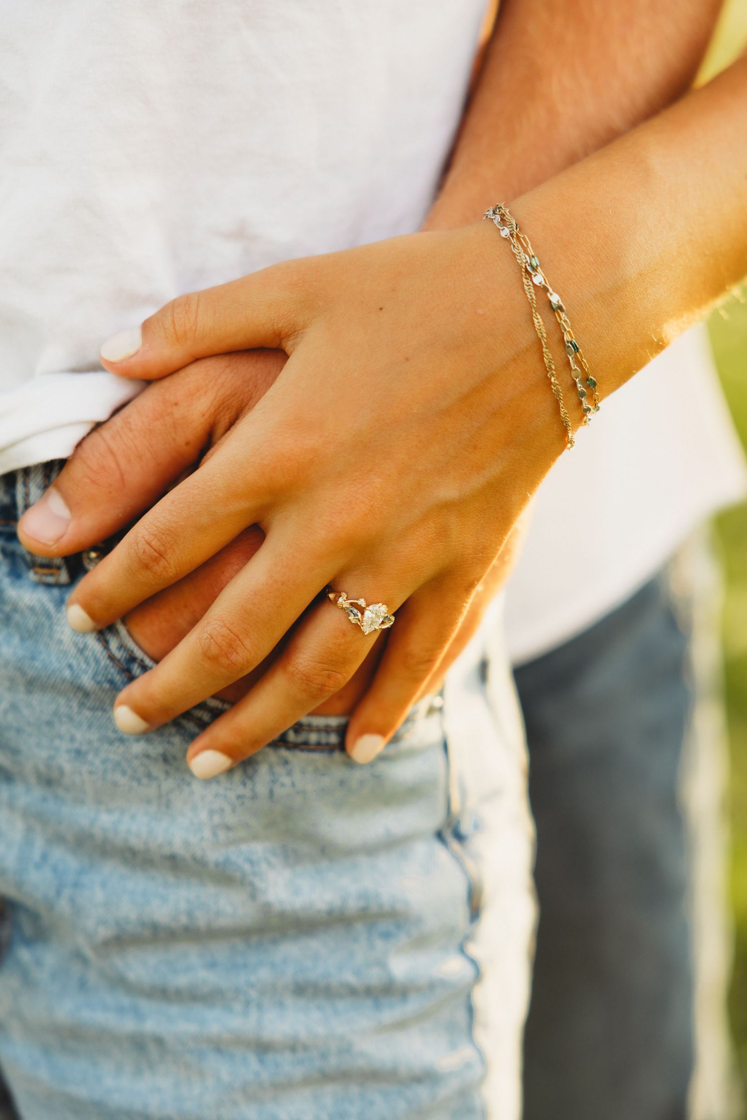 Close-up of a woman's hand wearing a diamond engagement ring and gold chain bracelet resting on a man's jeans pocket.