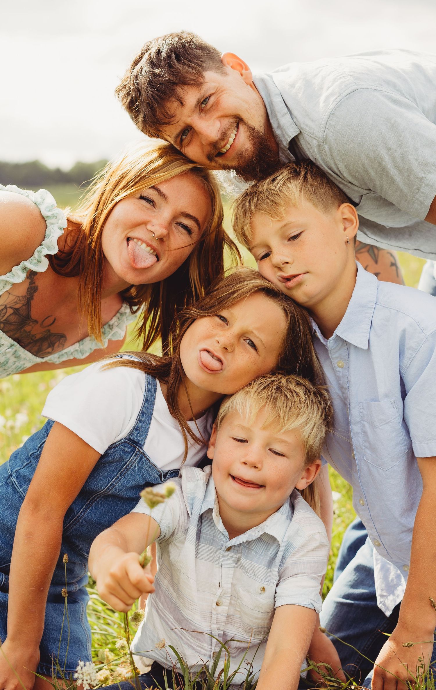 Smiling family of five with two adults and three children posing close together and sticking out tongues in a sunny flower field.