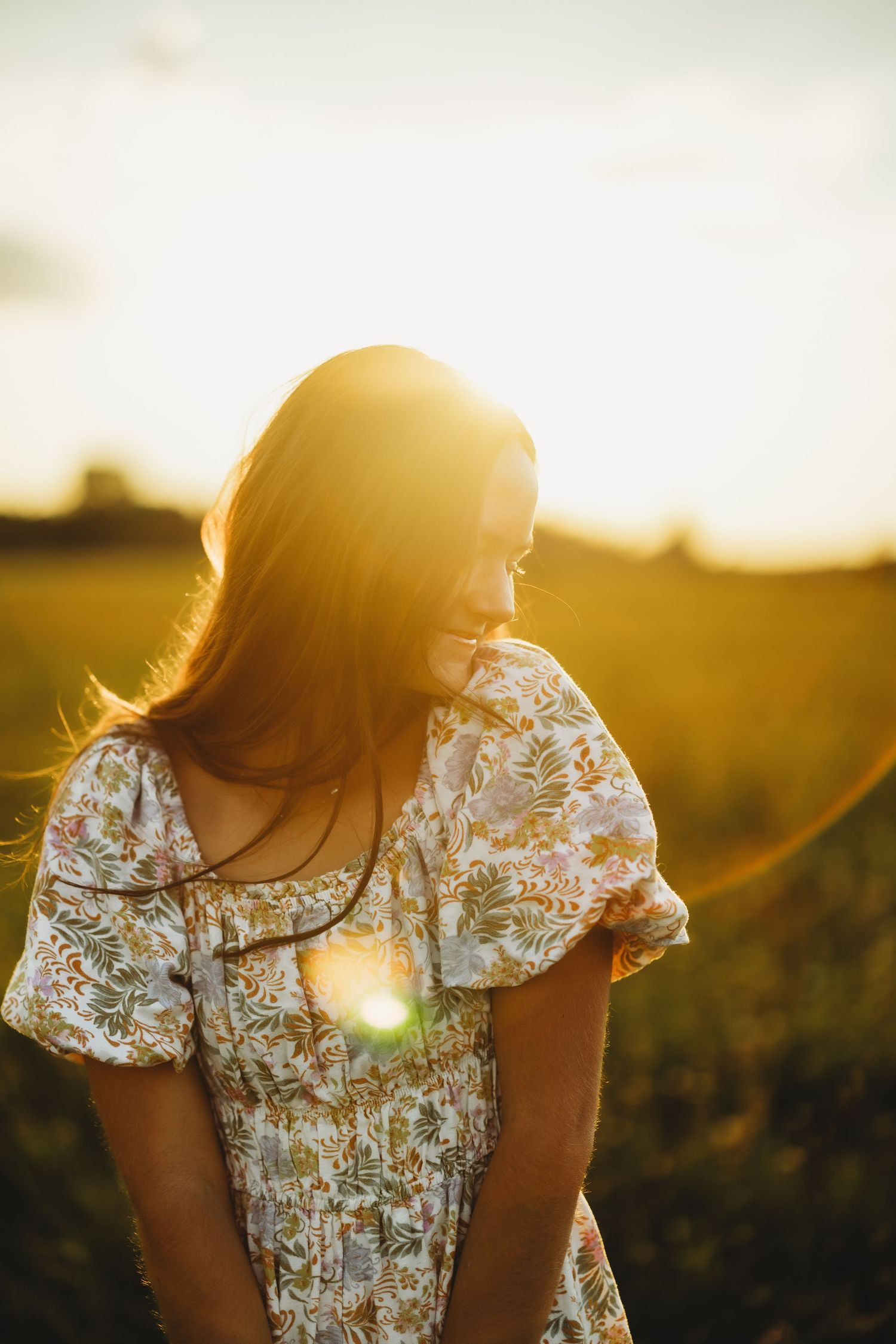 Woman in a floral dress glances down in a sunlit field as golden sunset light and lens flare glow behind her.