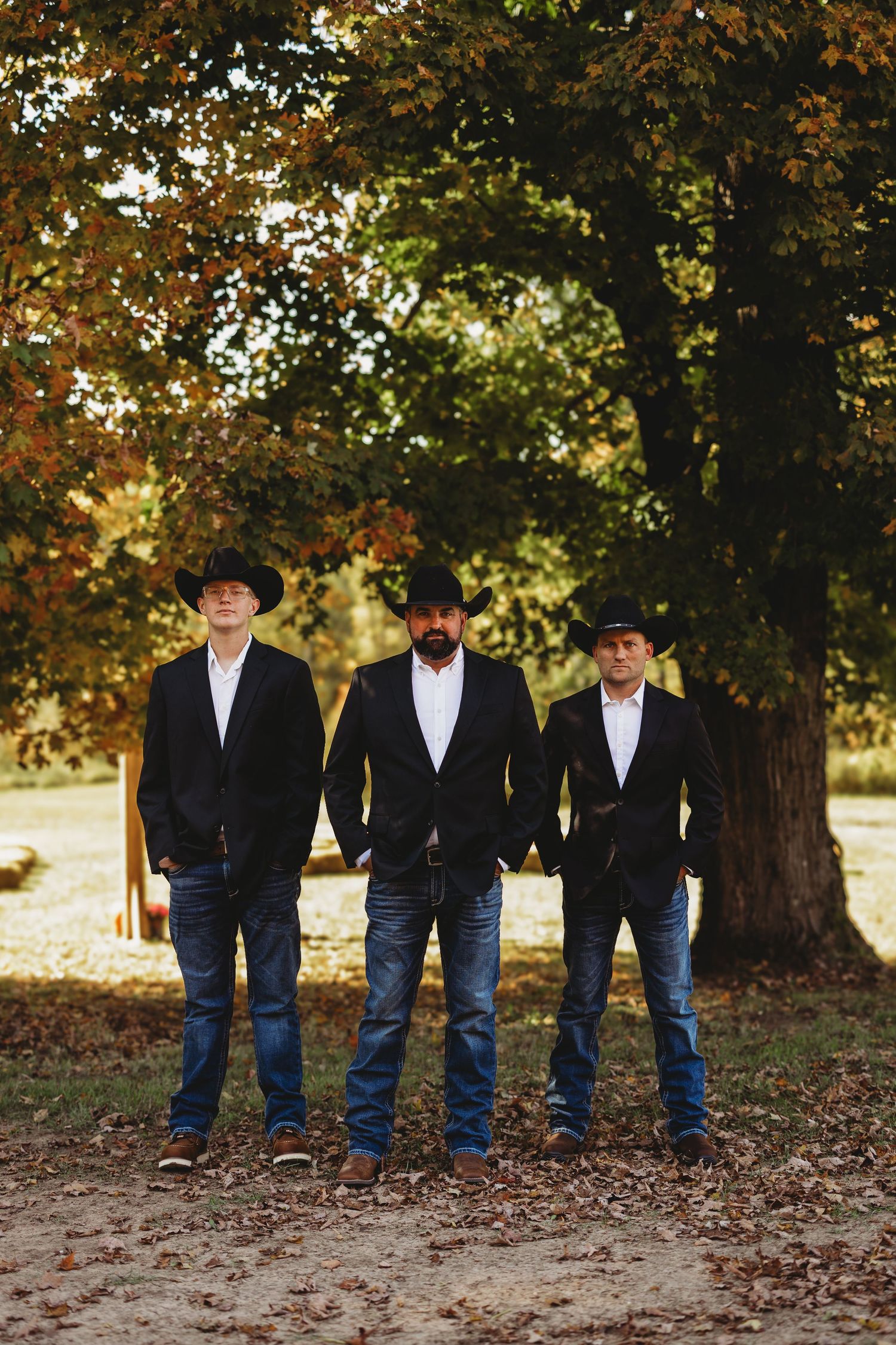 Three groomsmen in black jackets, jeans and cowboy hats stand side by side on a dirt path beneath autumn trees.