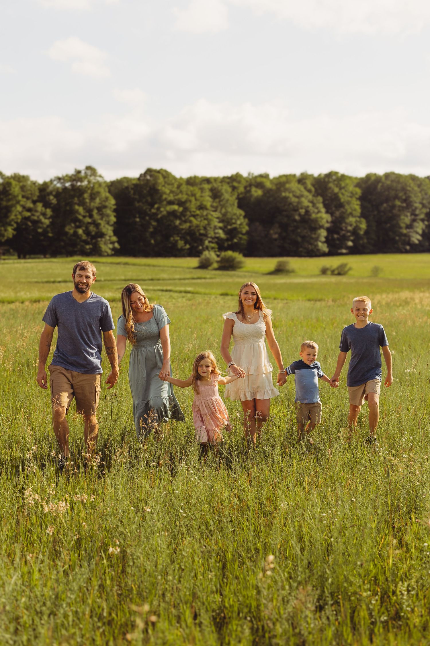 A happy family of six walks hand-in-hand through a sunny green meadow with trees in the background.