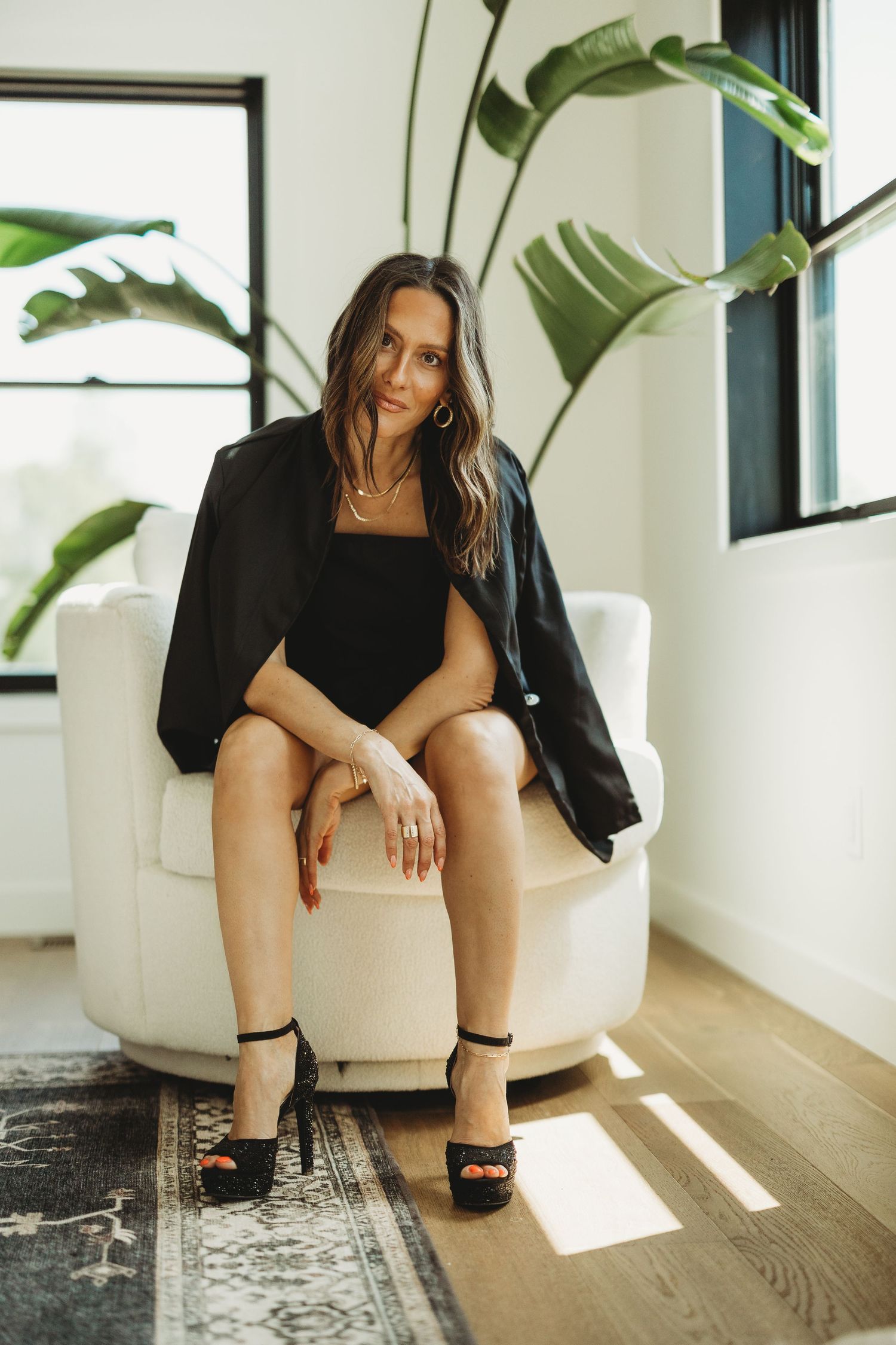Woman in black blazer and heels sitting elegantly in a white chair with tropical plant backdrop.