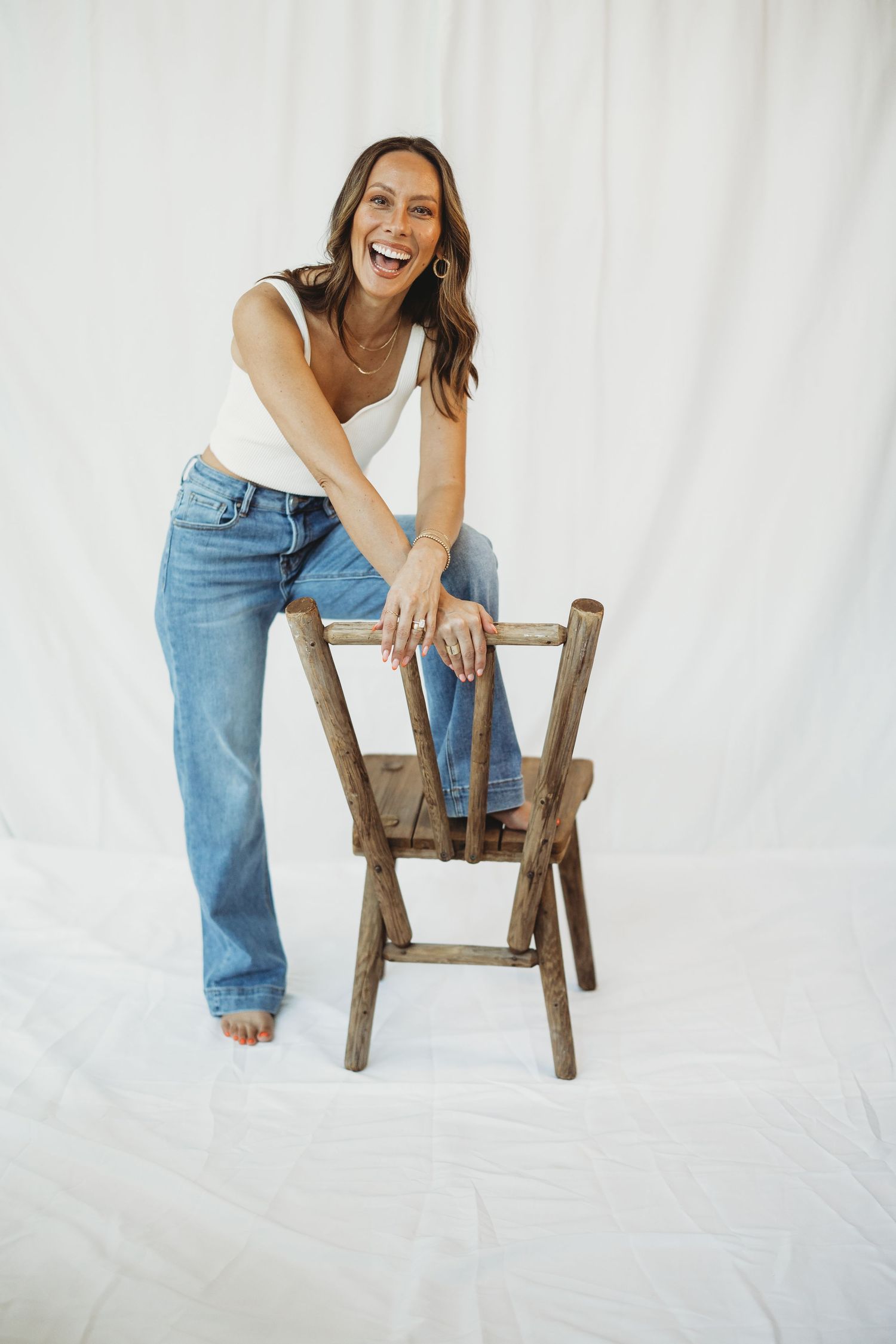 Laughing woman in white tank top and blue jeans leaning on rustic wooden chair against white backdrop.
