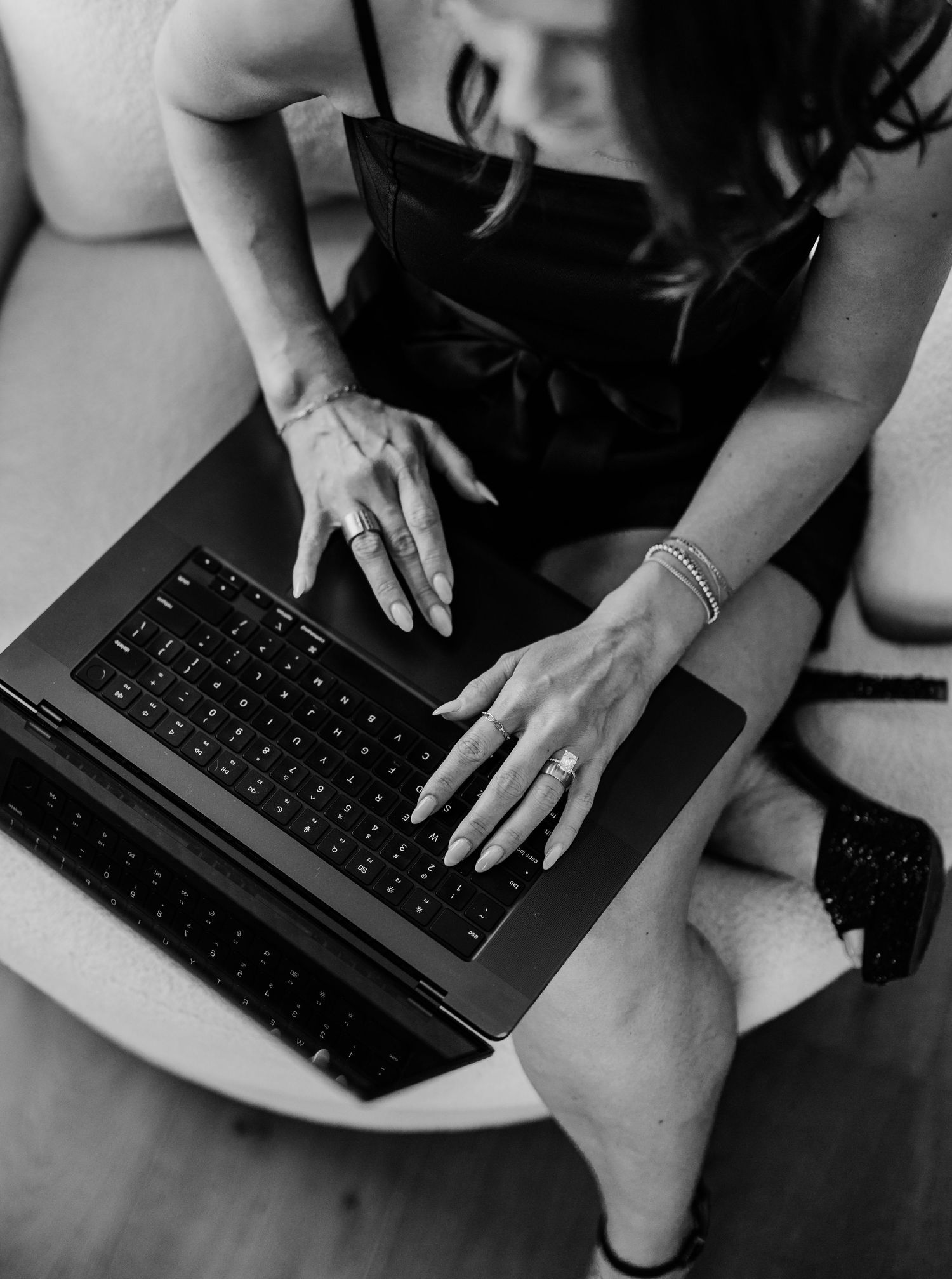 Black and white photo of a woman in a black dress typing on a laptop, wearing rings and bracelets.