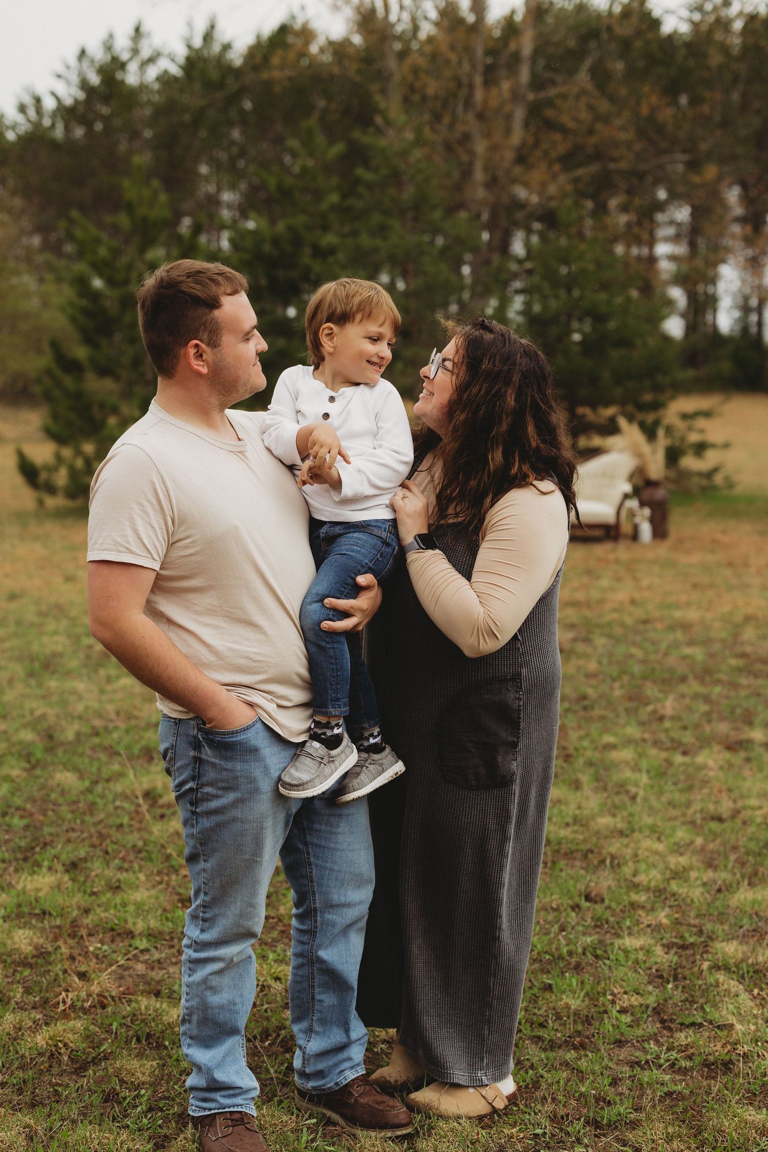 Family of three posing outdoors, father holding toddler son while mother smiles, surrounded by trees in a field.