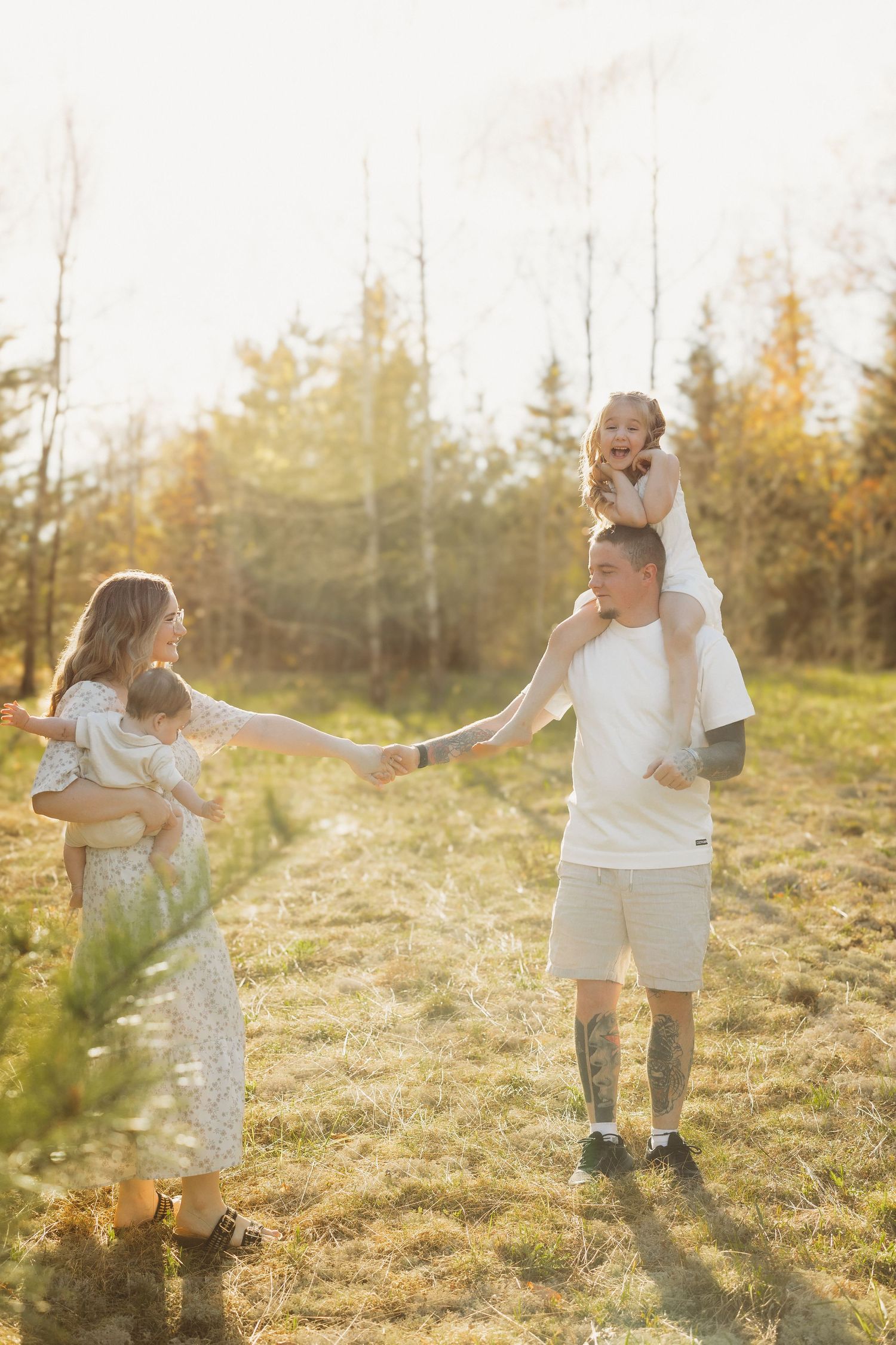 Happy family holding hands in golden sunlit field, mother carrying baby, father with toddler on shoulders.