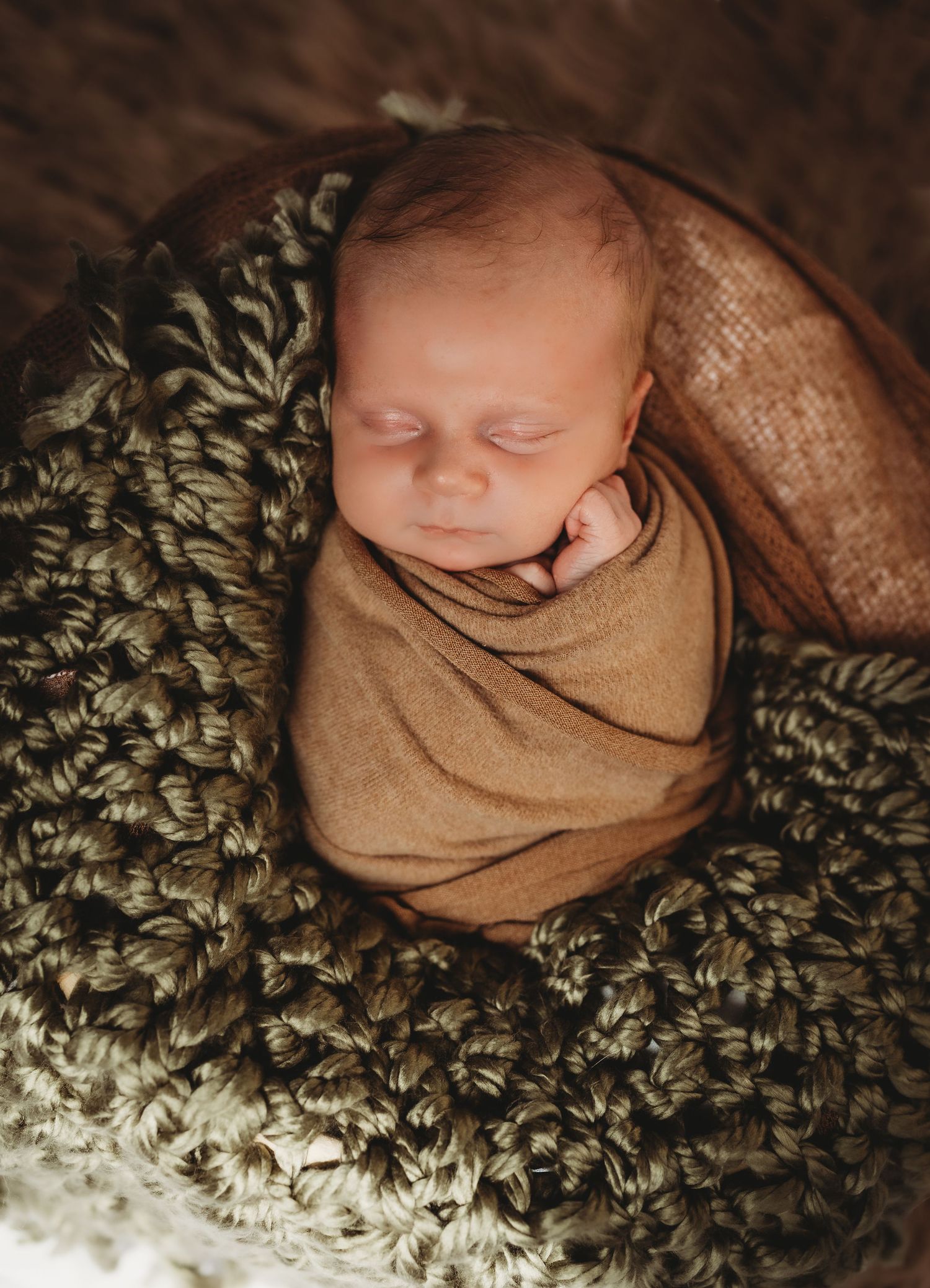 Sleeping newborn wrapped in brown swaddle lying on textured green blanket.