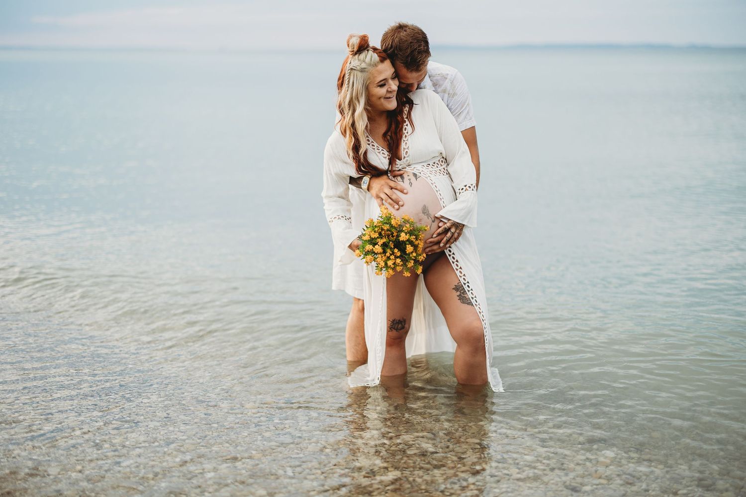 Romantic couple standing in shallow ocean water with yellow bouquet during beach photoshoot.