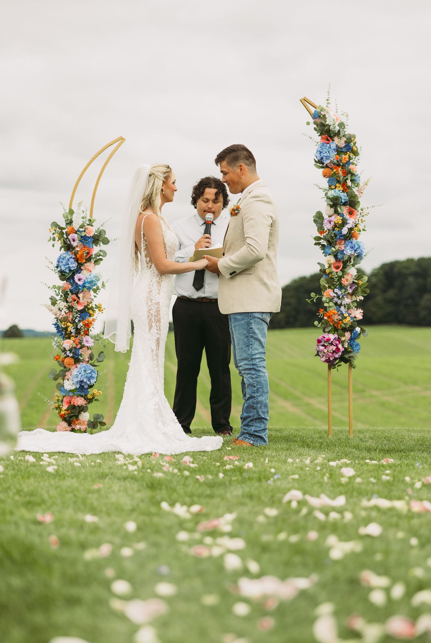 Wedding ceremony takes place outdoors with colorful floral arch and scattered petals on grassy field.