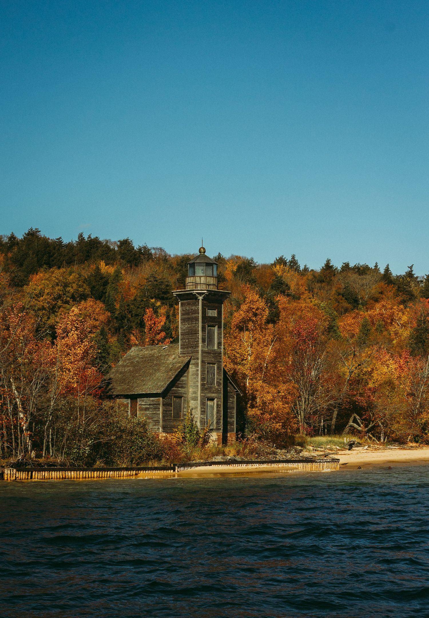 Historic wooden lighthouse stands among autumn trees on Lake Superior shoreline at sunset.