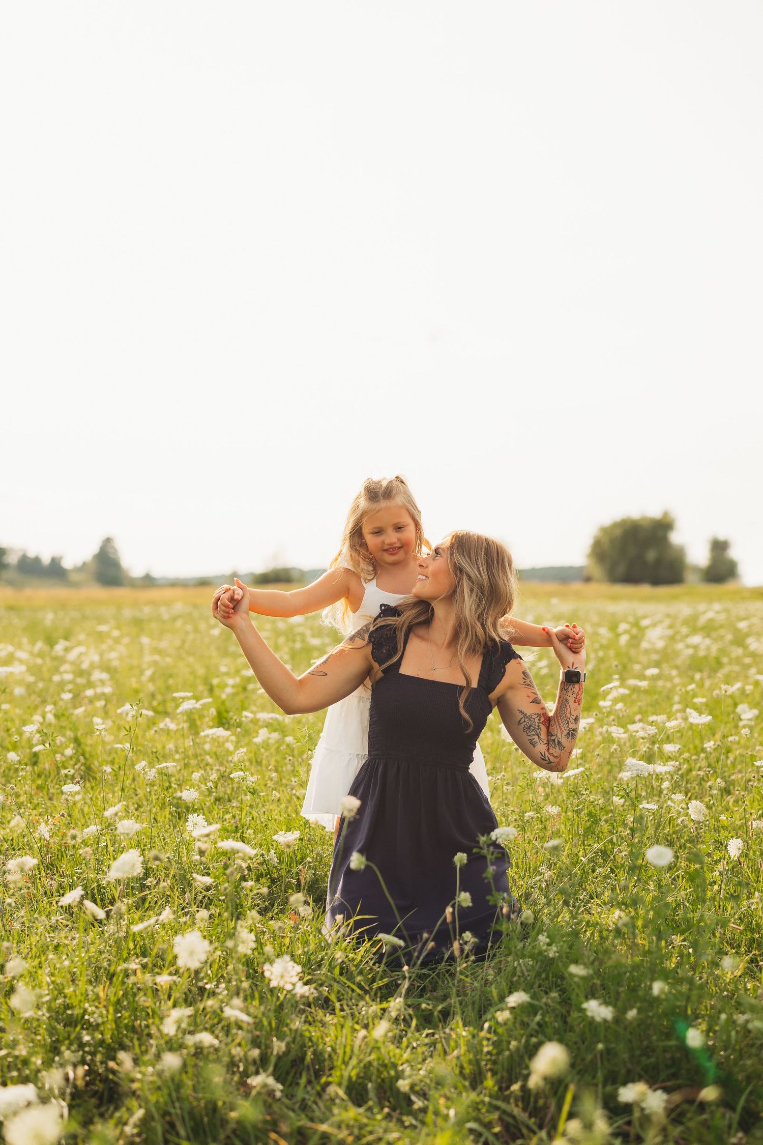 A tattooed mother lifts her smiling daughter in a sunny wildflower field, both sharing a joyful moment outdoors.