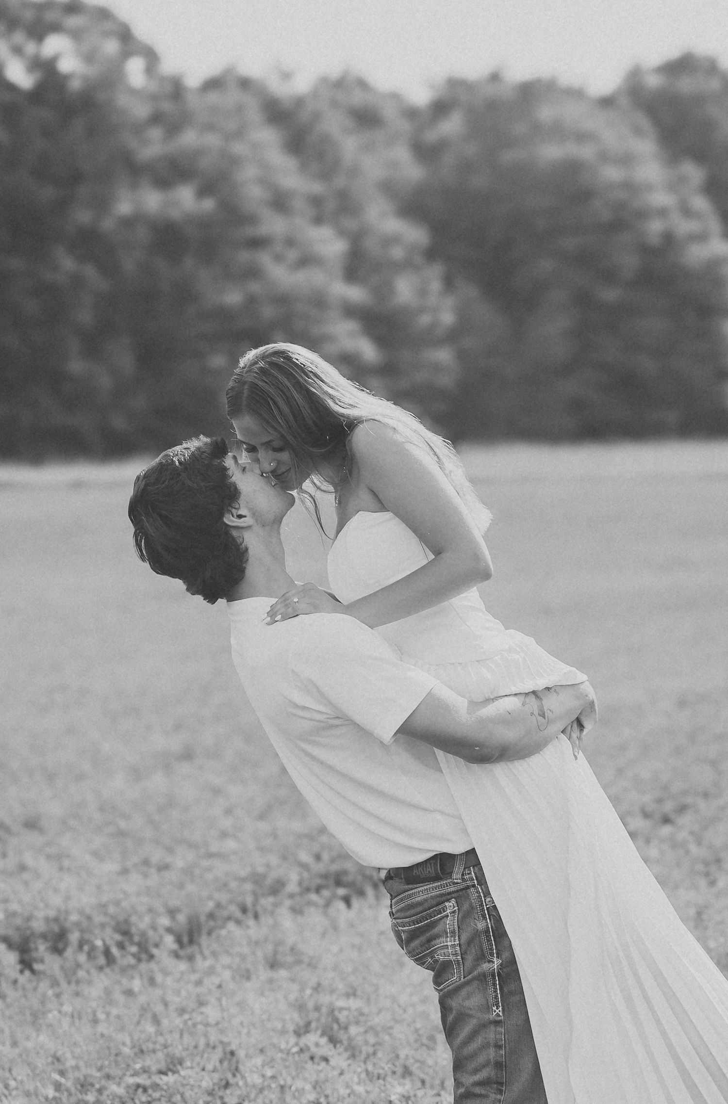 Black and white photo of a man lifting and kissing a woman in a white dress in an open field.