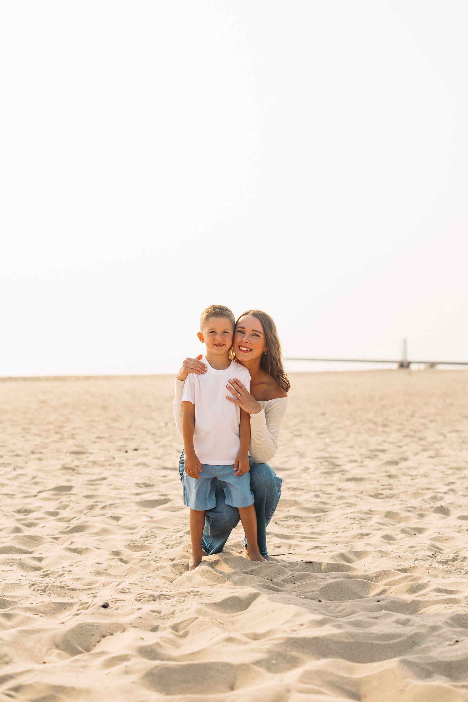 A mother kneels on sandy beach hugging her young son, both smiling in casual white tops and jeans.