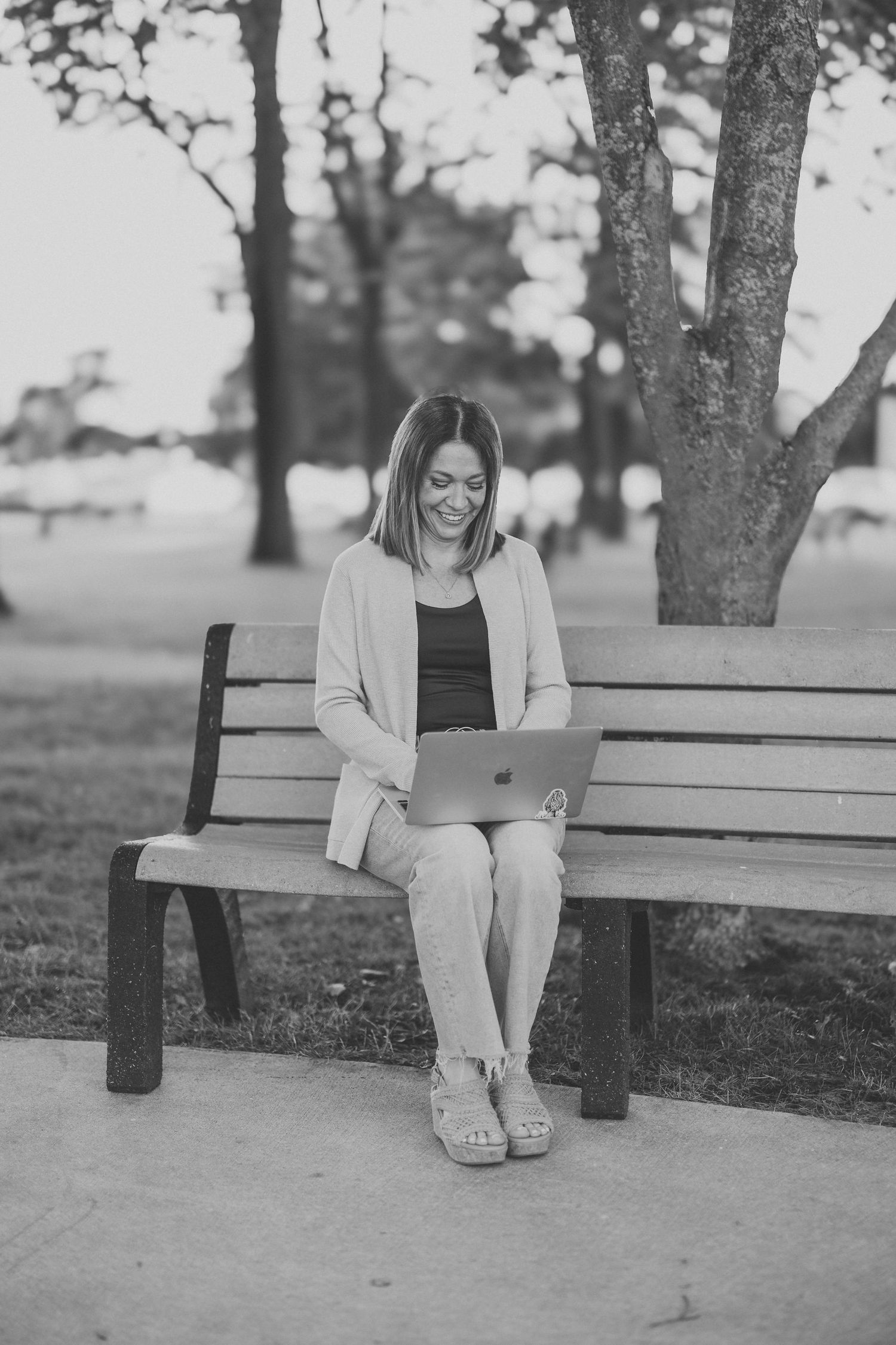 Woman smiling while using a laptop on a park bench, surrounded by trees in a serene outdoor setting.