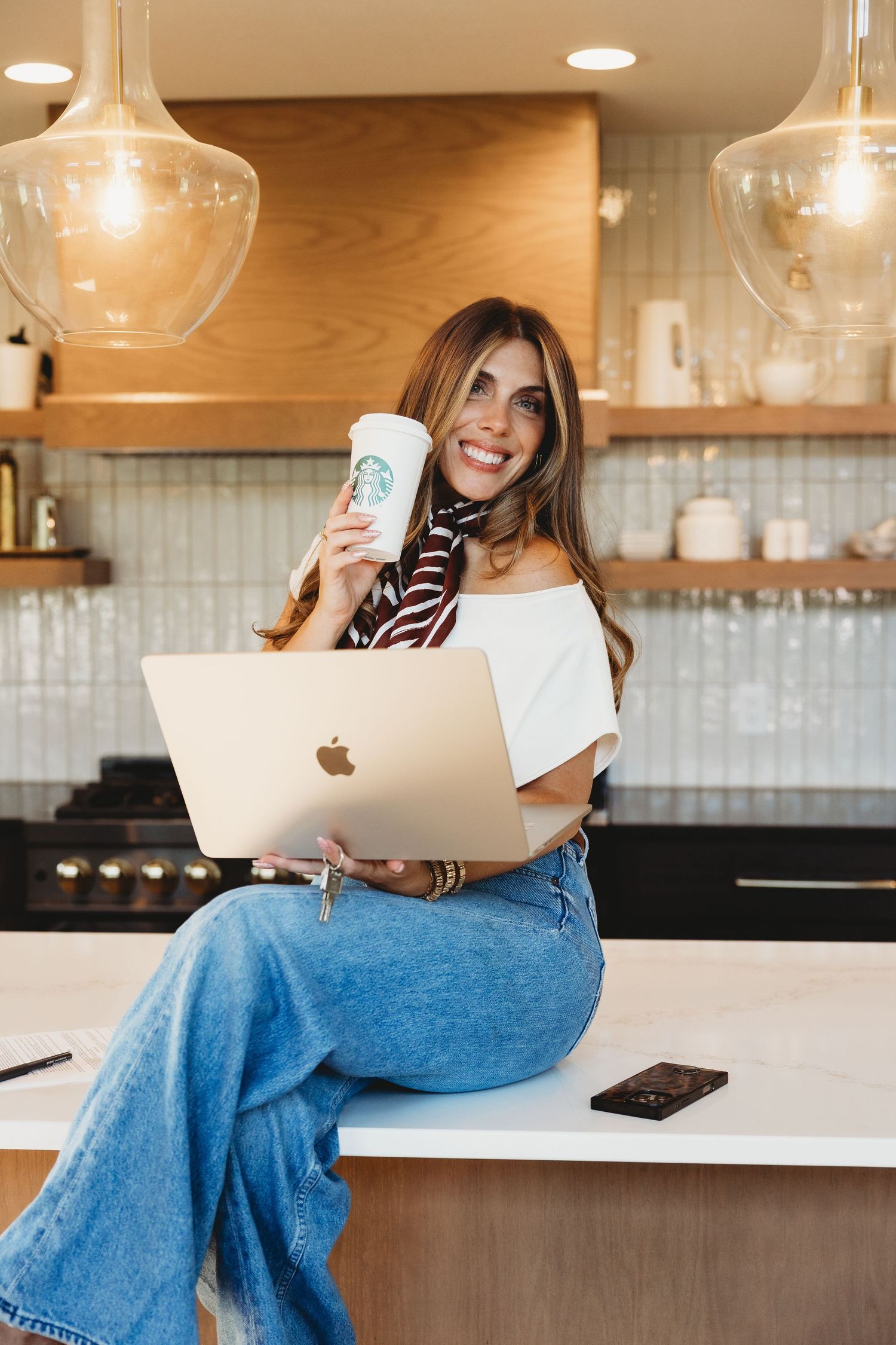 Woman in jeans and a striped scarf sits on a kitchen counter holding a Starbucks cup and an open MacBook laptop.