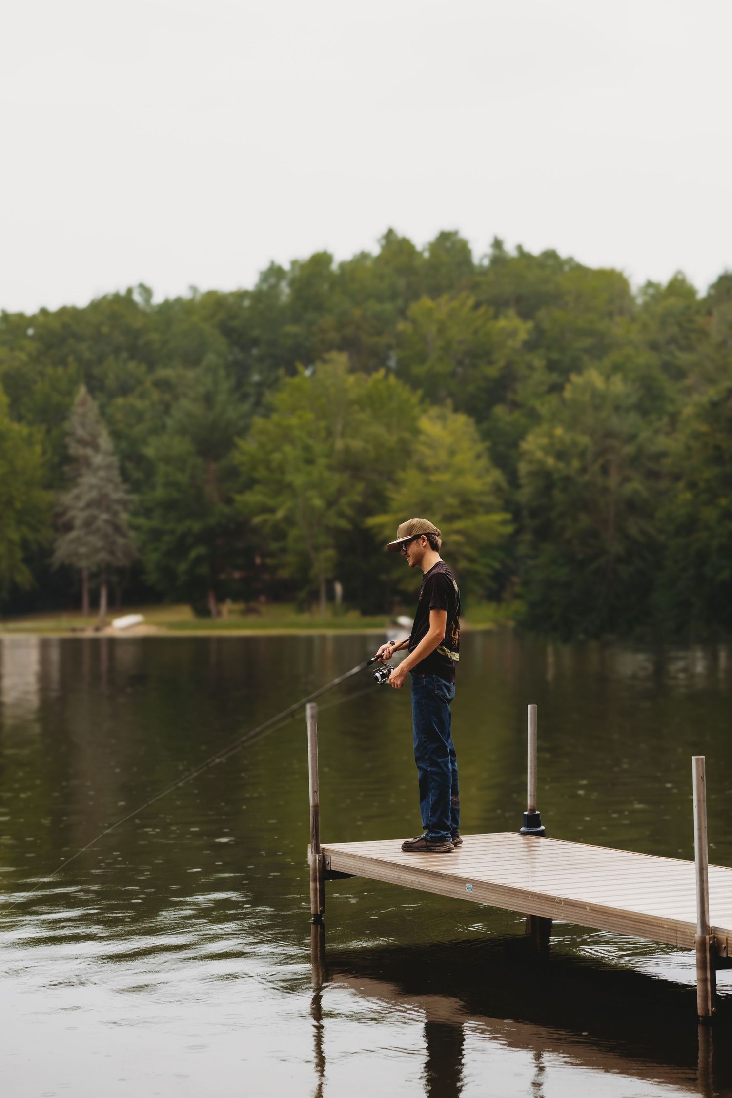Angler in a cap and jeans stands on a wooden lake dock casting a fishing rod over calm water, with green forest across the shore.