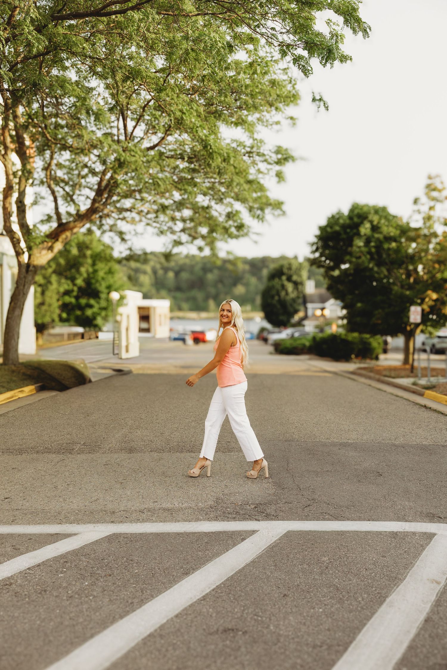 Woman in a pink top and white pants walks across a crosswalk on a quiet tree-lined street, smiling back at the camera on a sunny evening.