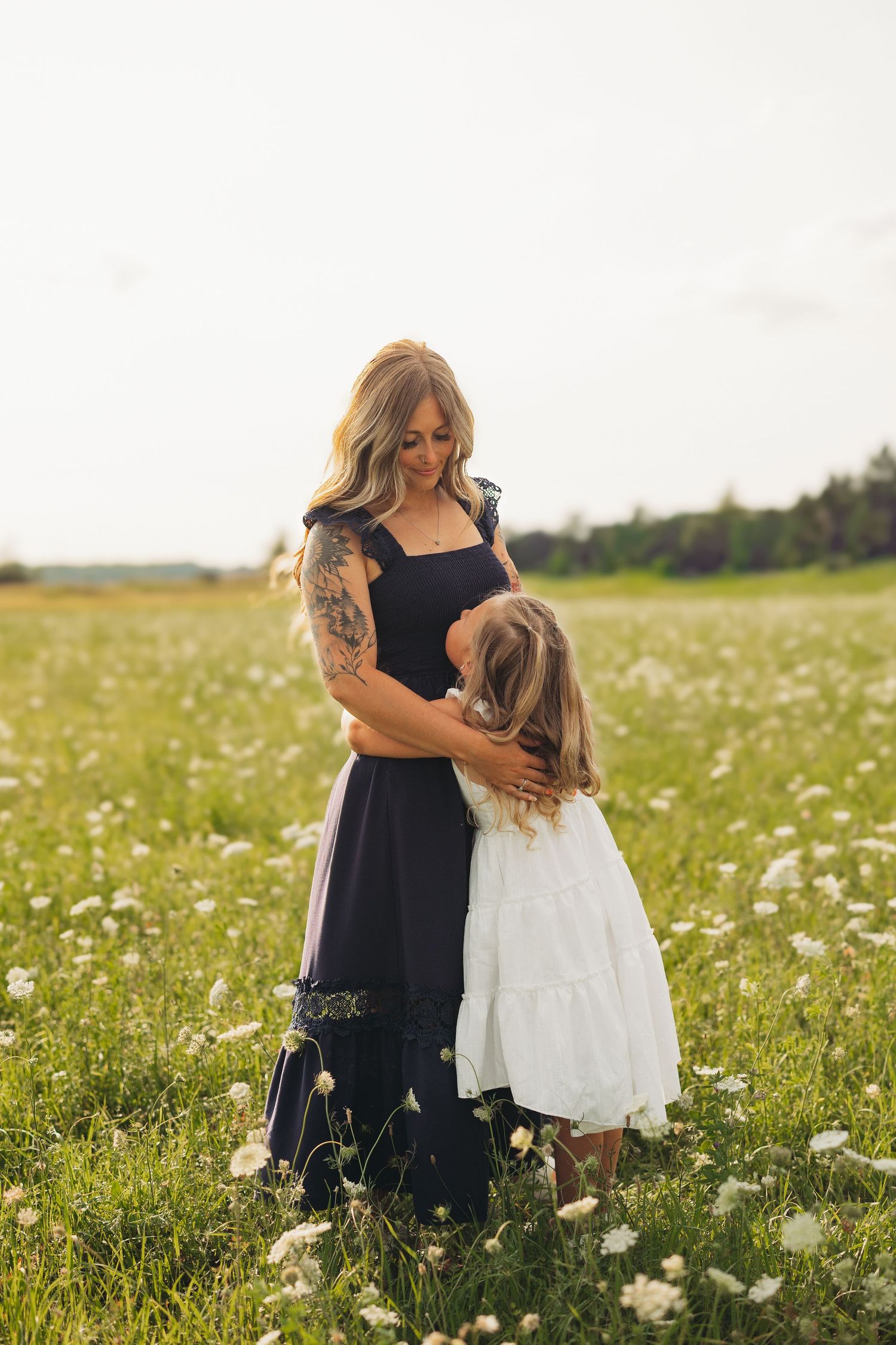 Mother in a navy dress embracing her young daughter in a white dress in a sunlit meadow of white wildflowers at golden hour.