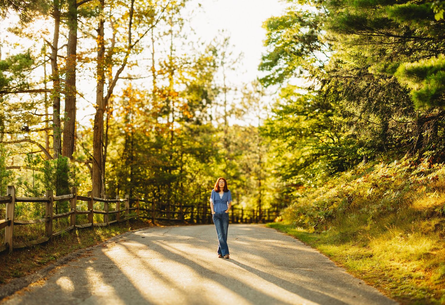 Woman in a blue top and jeans walks down a sunlit paved path lined with a wooden fence and golden autumn trees.
