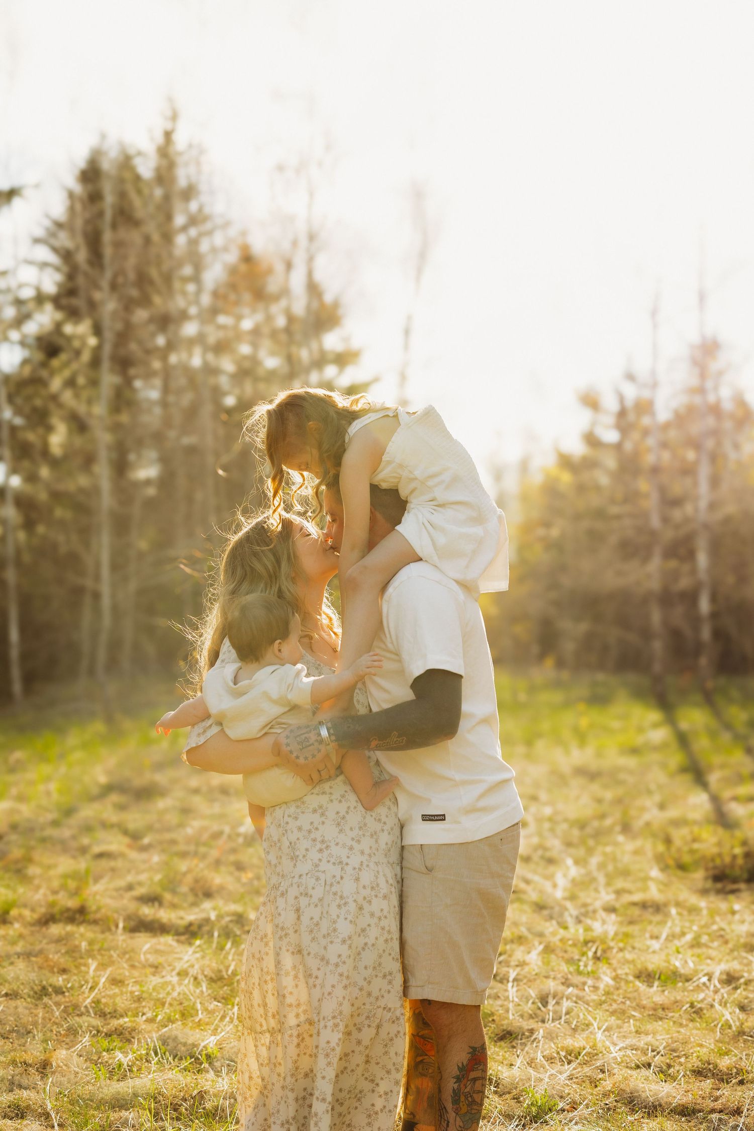 A family shares a warm embrace in a sunlit field, with a child lifted up and trees glowing in the background.