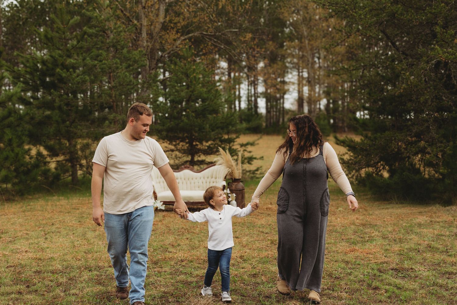 Family of three walking hand-in-hand through a green meadow with pine trees, parents swinging toddler between them.