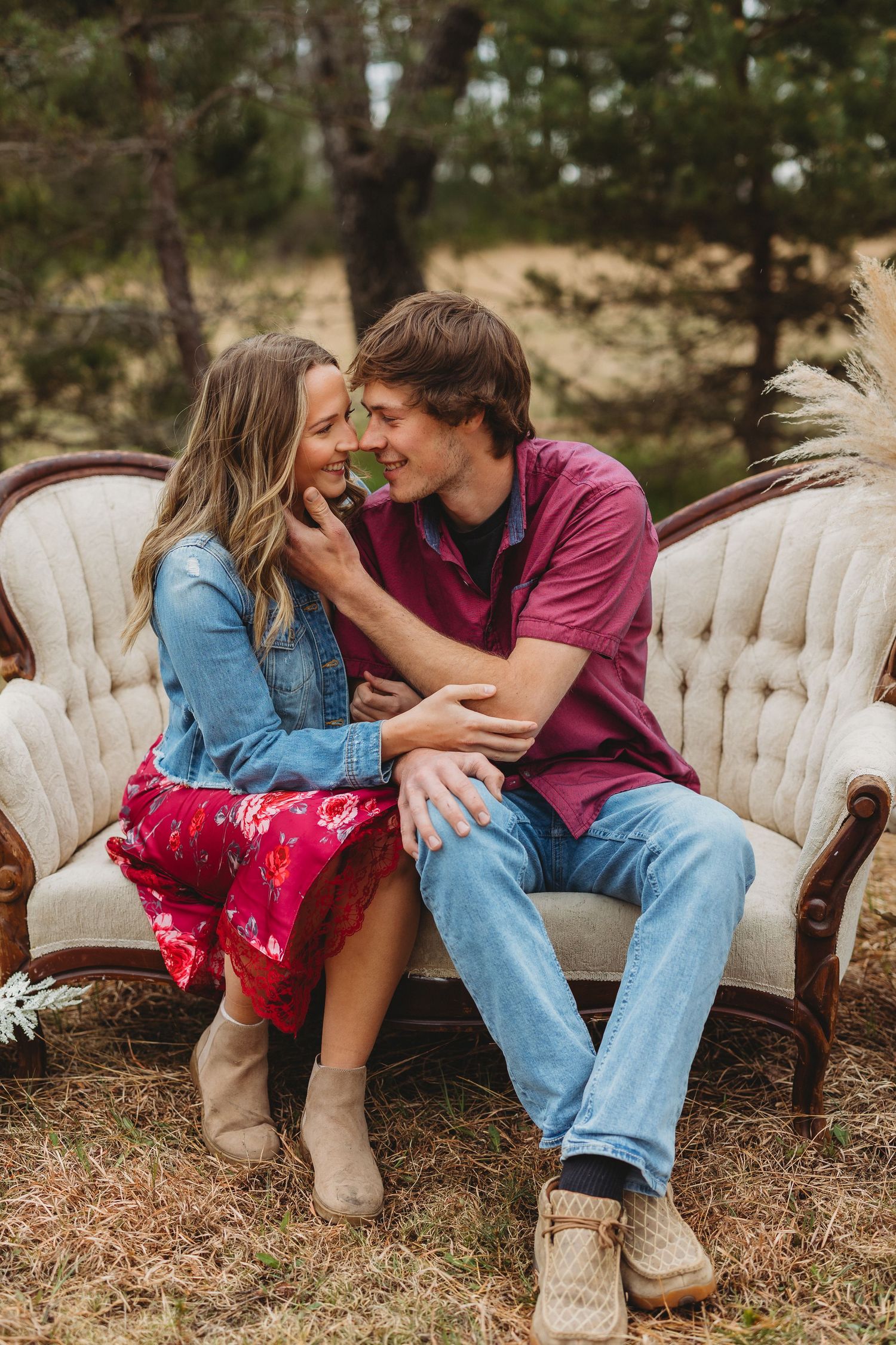 Couple sitting on vintage sofa outdoors, smiling at each other, woman in red floral skirt and denim jacket, man in burgundy shirt.