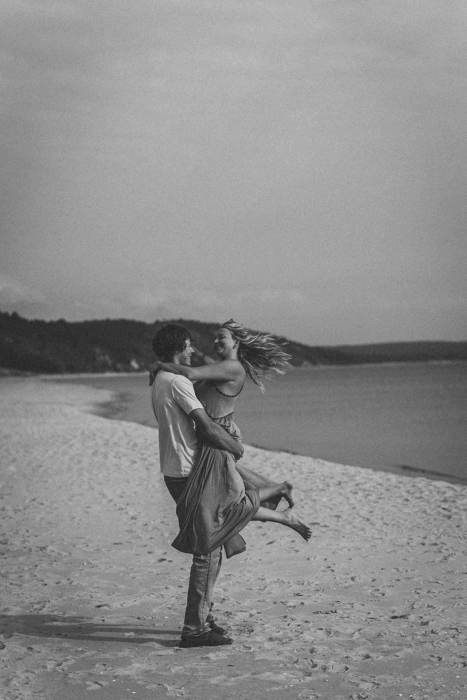 Black and white photos of a playful couple dancing and twirling on a sandy beach.