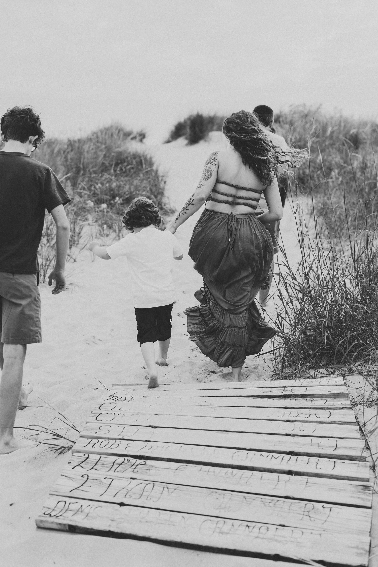 A black and white image of people walking on a wooden boardwalk through beach dunes.
