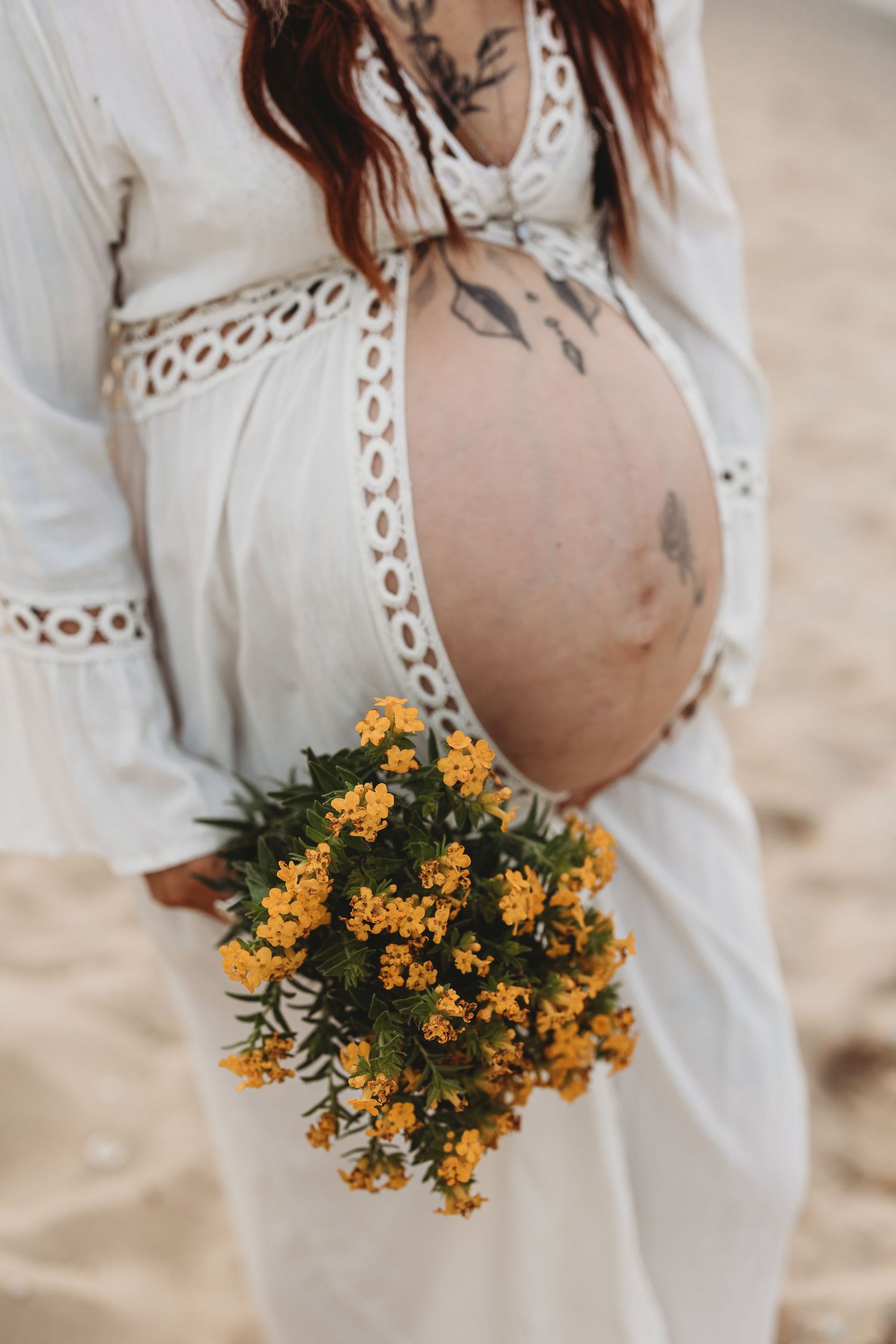 Pregnant woman in white lace dress holding yellow wildflowers showing baby bump.