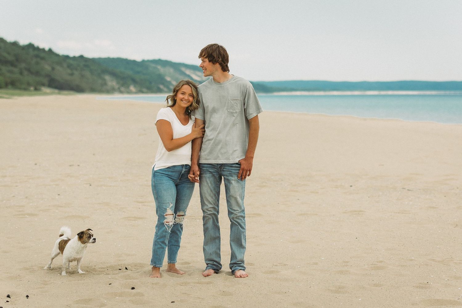 A couple walking along a beach with their small dog beside them.