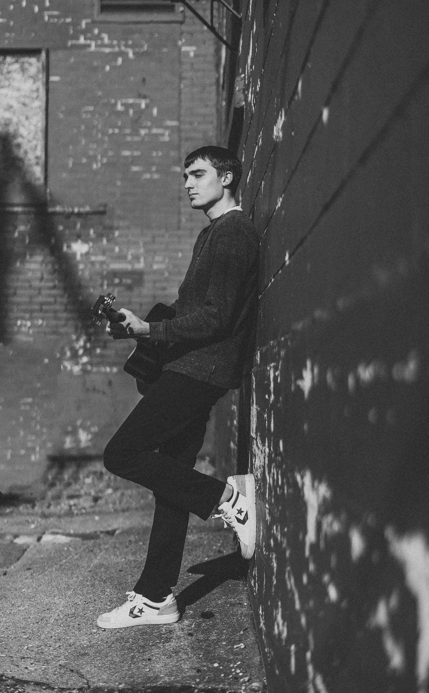 A moody portrait of a person with a guitar leaning against a brick wall.