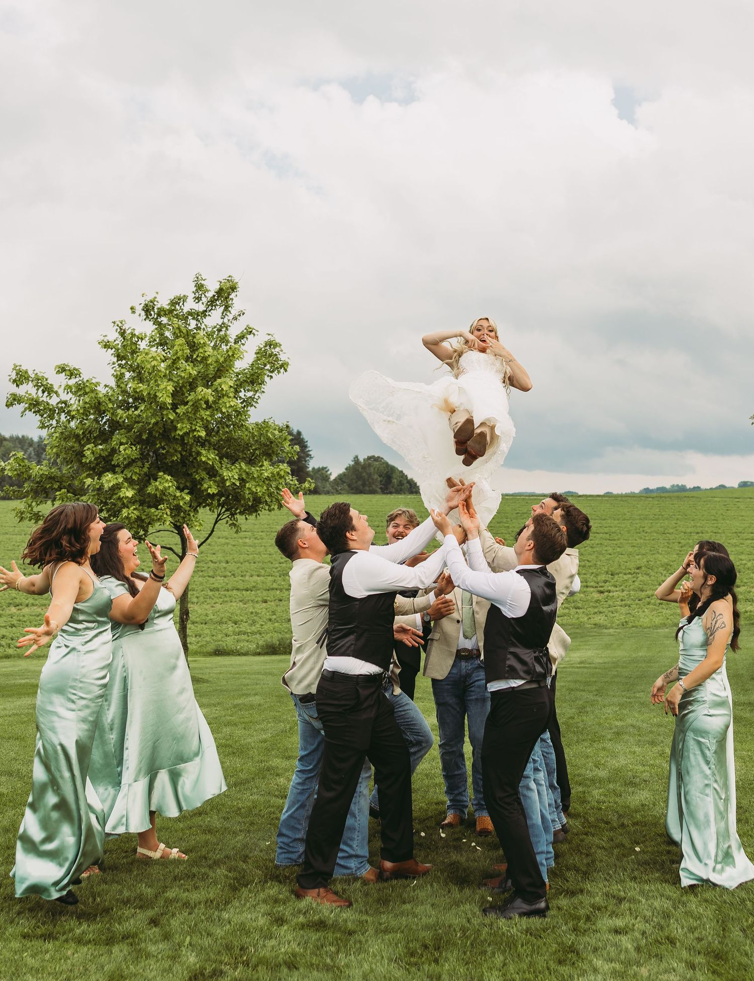 Wedding party members playfully toss flower petals into the air while standing in a grassy field on a cloudy day.