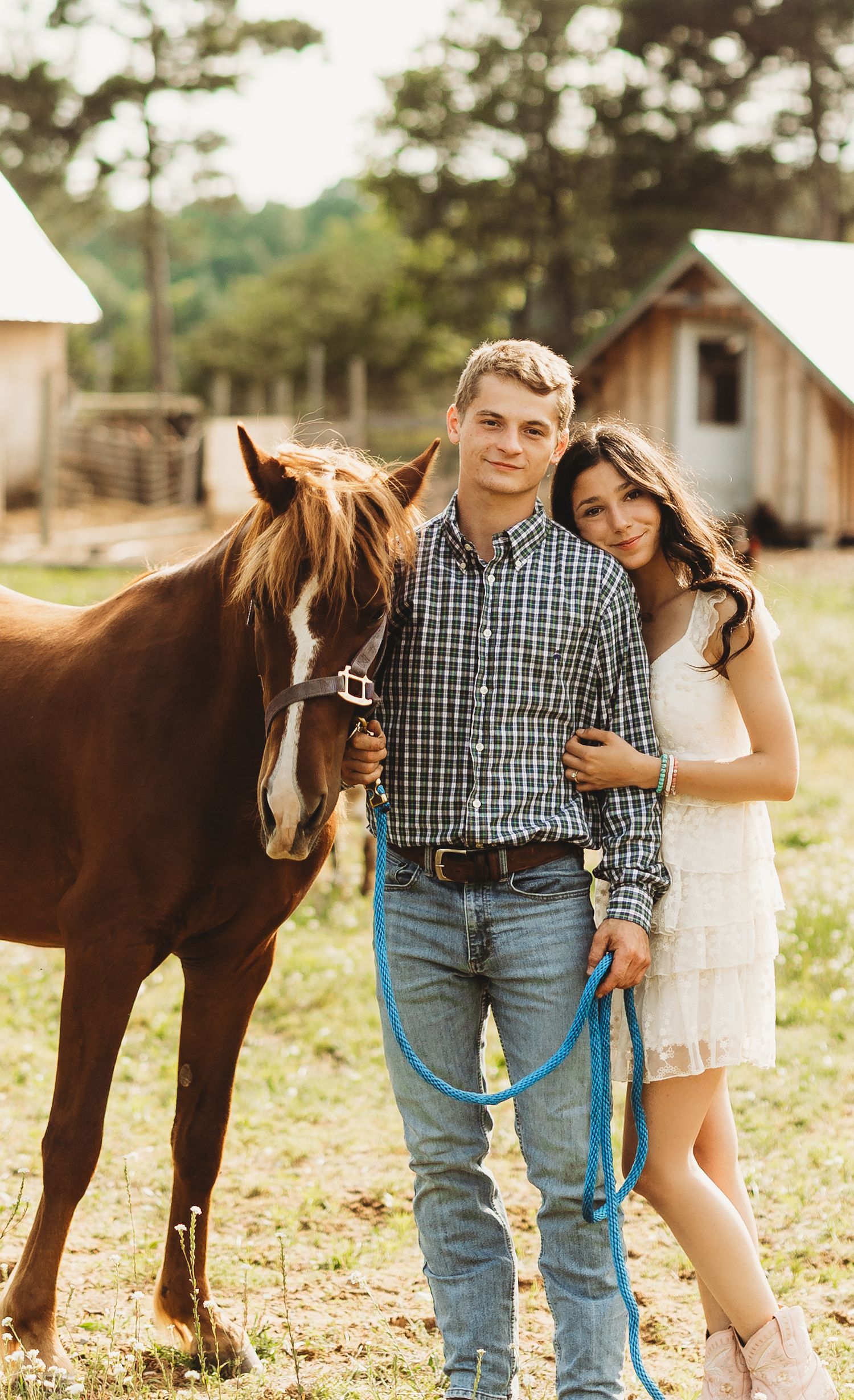 A couple stands with their brown horse on a sunny day at a rustic farm with wooden cabins in the background.