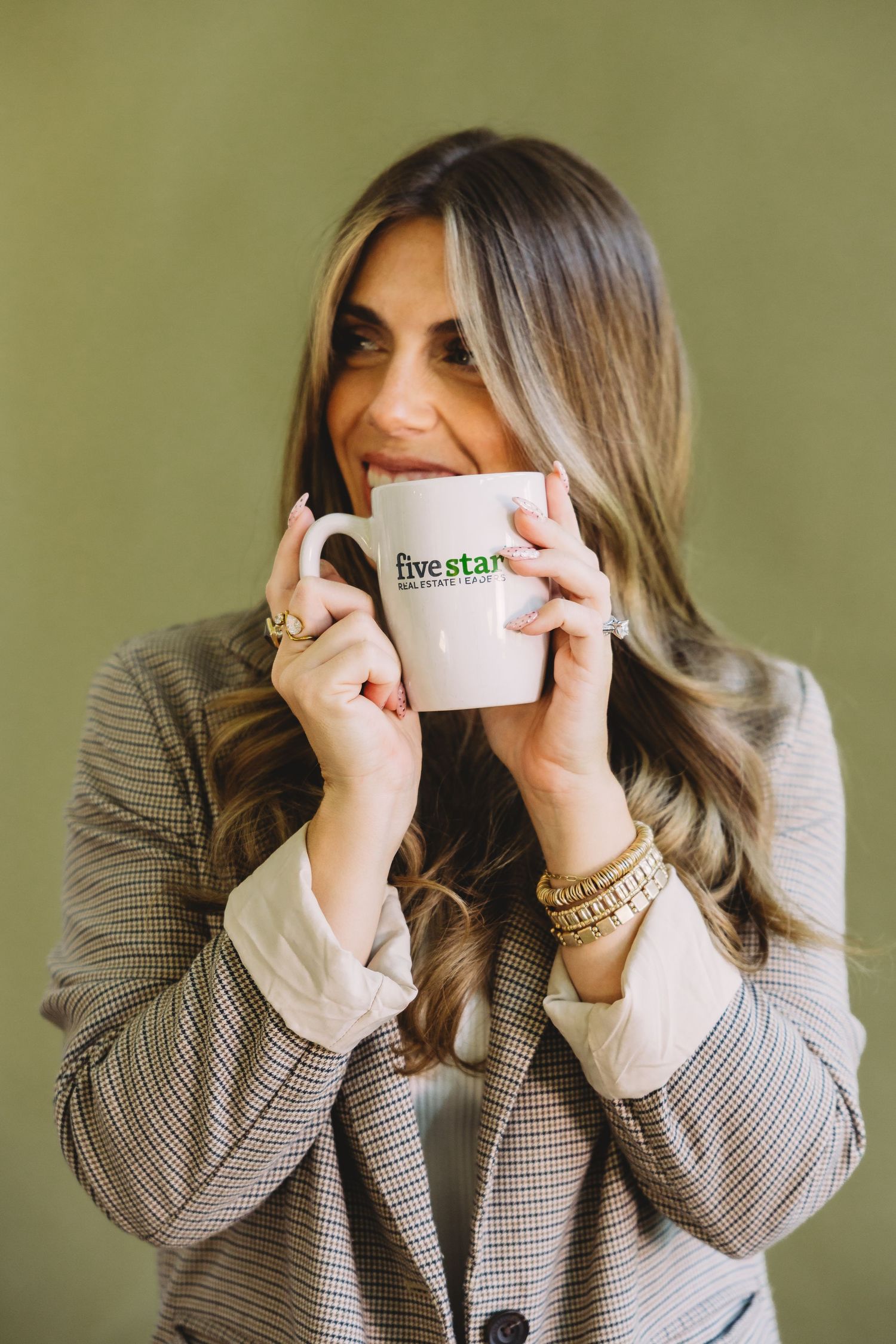 Woman with long highlighted hair in a plaid blazer sips from a white Five Star mug against an olive green wall.
