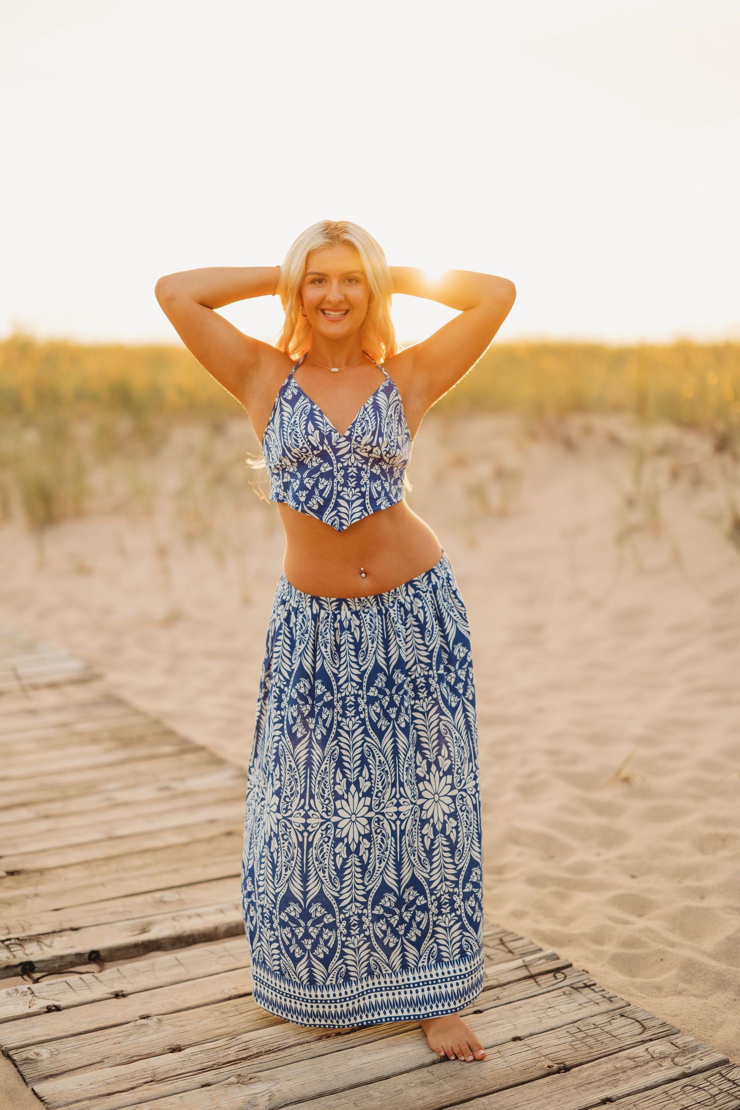Smiling blonde woman in a blue and white patterned crop top and maxi skirt posing with hands behind her head on a beach boardwalk.